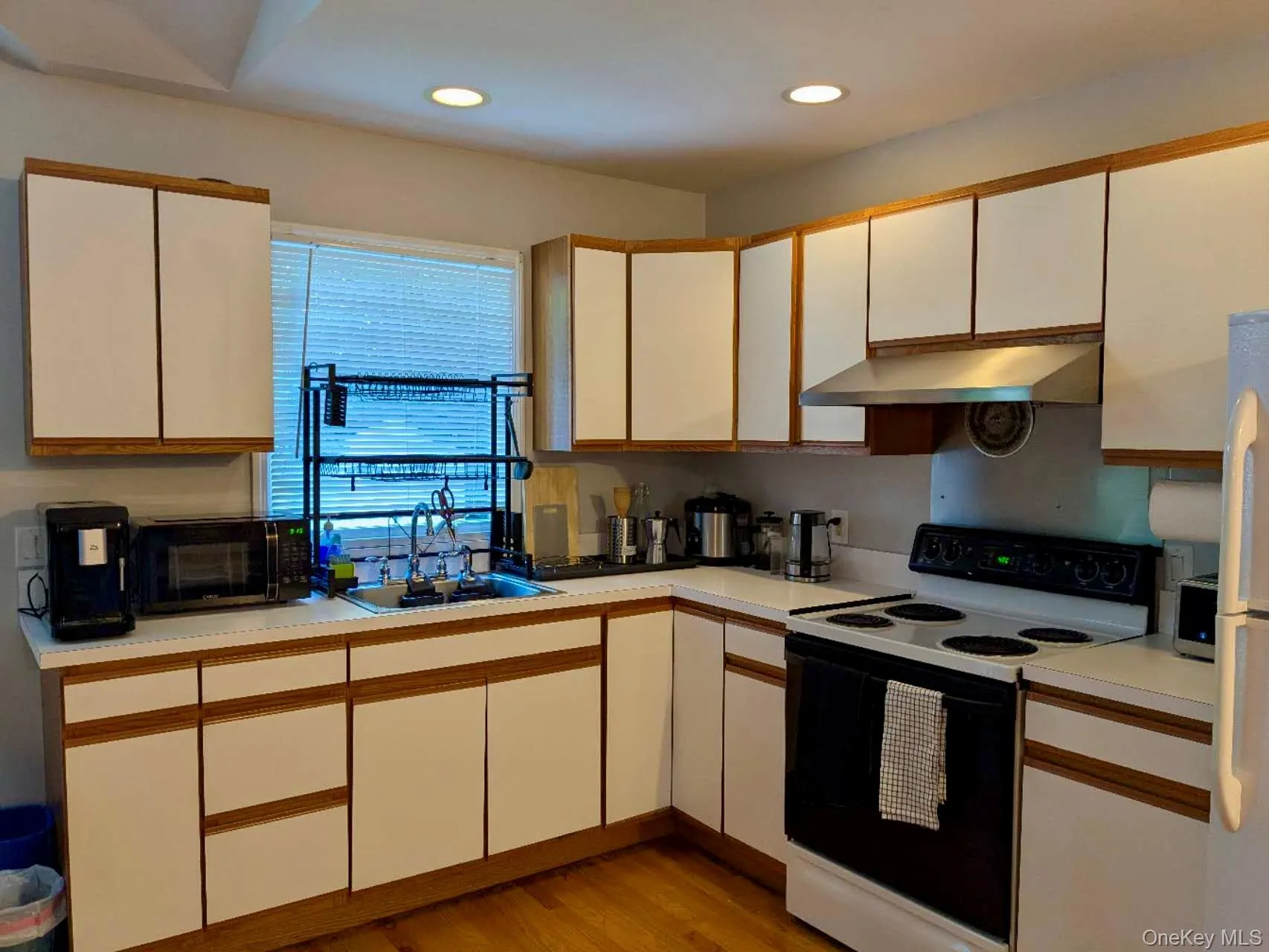 Kitchen featuring a white appliance package, light wood-finish flooring, recessed lighting, white cabinetry with wood trim, and a stainless steel range hood Kitchen featuring a white appliance package, light wood-finish flooring, recessed lighting, white cabinetry with wood trim, and a stainless steel range hood