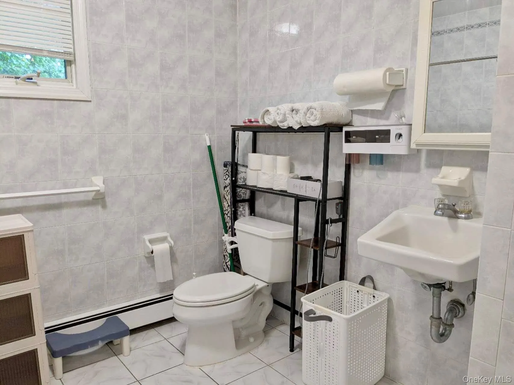 Bathroom featuring floor-to-ceiling tile, a single vanity with an integrated sink, a toilet, a window with blinds, and a wall-mounted mirror Bathroom featuring floor-to-ceiling tile, a single vanity with an integrated sink, a toilet, a window with blinds, and a wall-mounted mirror