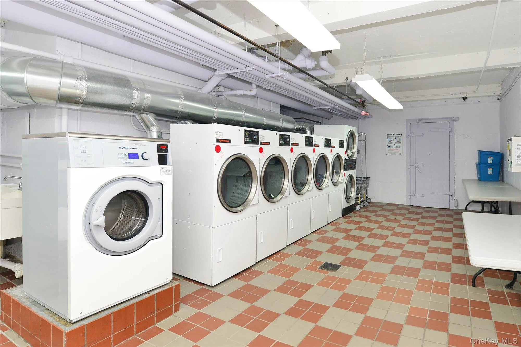 Spacious laundry room with a checkerboard tile floor, multiple commercial-grade washers and dryers, exposed overhead ductwork, and fluorescent lighting Spacious laundry room with a checkerboard tile floor, multiple commercial-grade washers and dryers, exposed overhead ductwork, and fluorescent lighting