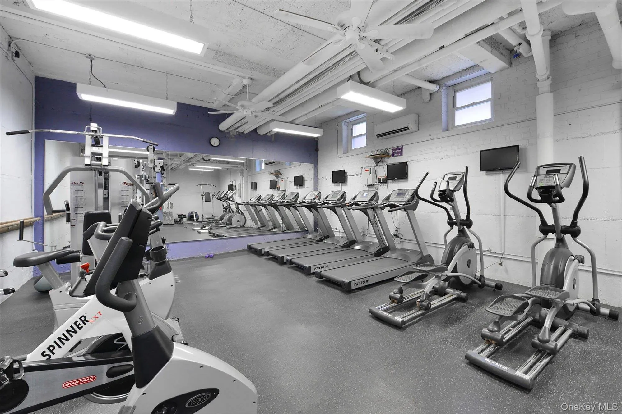 Fitness room featuring resilient flooring, mirrored wall, multiple cardio machines, exposed ceiling ductwork, and fluorescent lighting Fitness room featuring resilient flooring, mirrored wall, multiple cardio machines, exposed ceiling ductwork, and fluorescent lighting