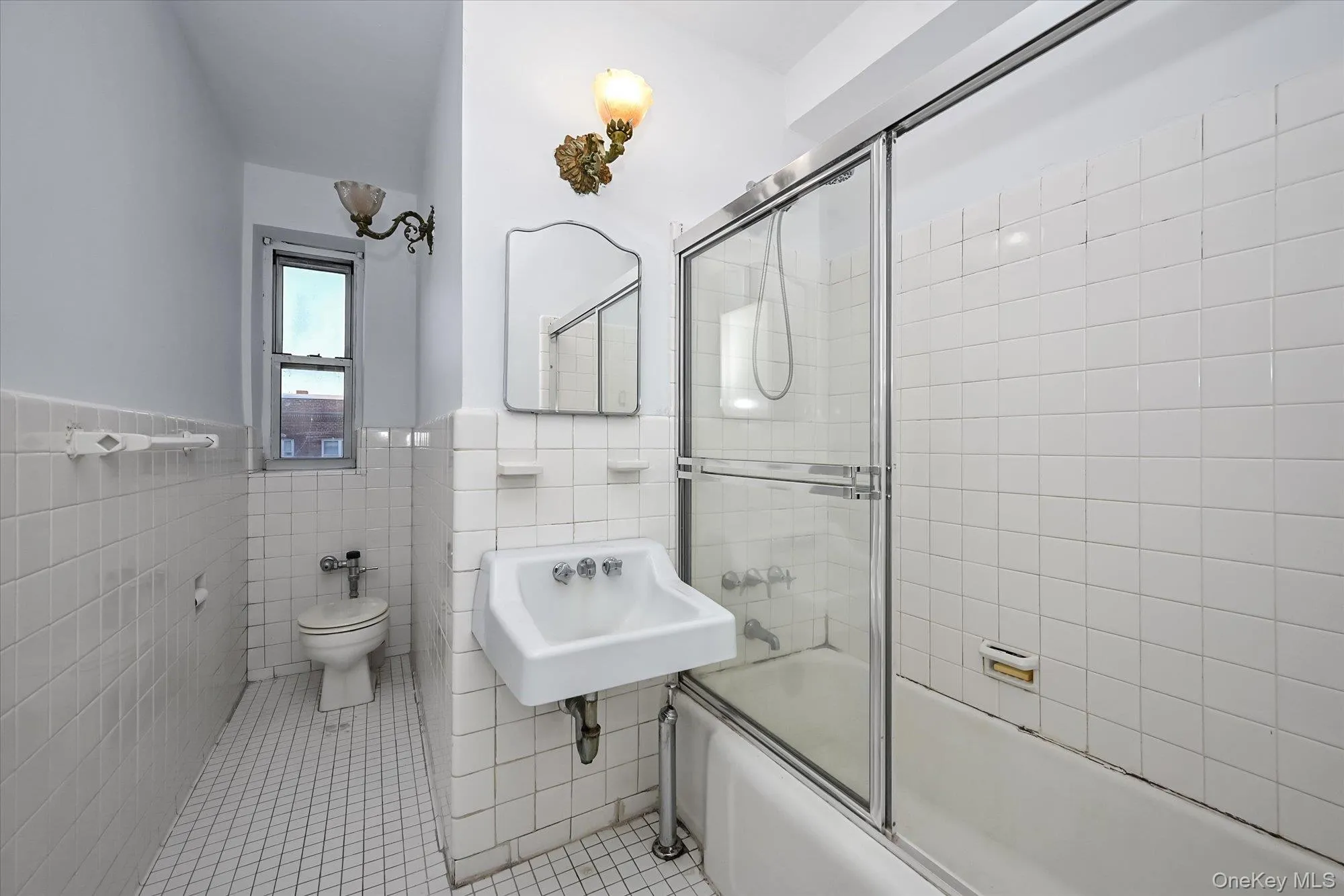 White tile walls and flooring feature in this bathroom, complete with a wall-mounted sink, toilet, and bathtub with glass enclosure White tile walls and flooring feature in this bathroom, complete with a wall-mounted sink, toilet, and bathtub with glass enclosure