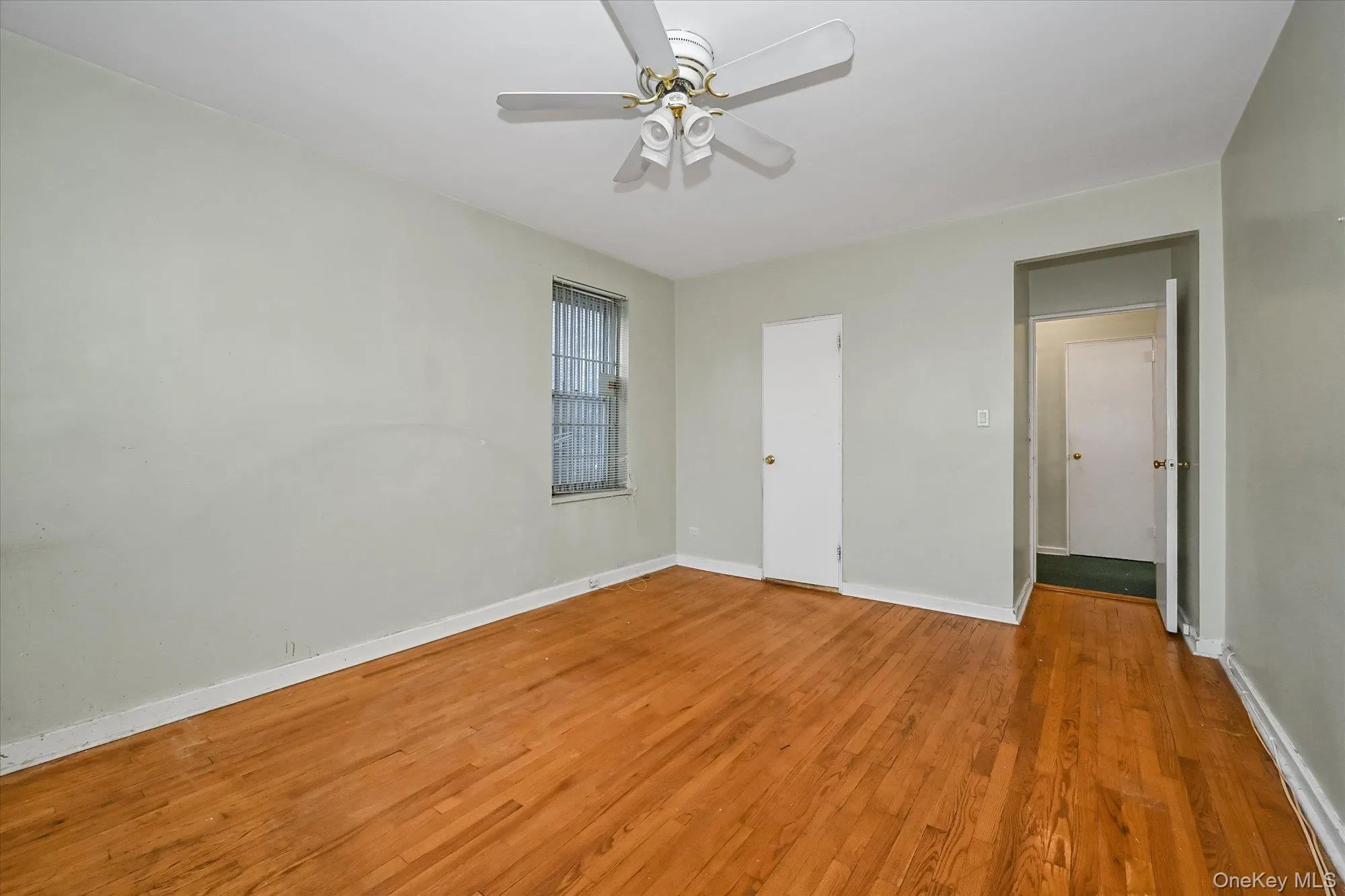 Spacious room featuring wood-finish flooring, light-colored walls, a ceiling fan fixture, and a window with security bars Spacious room featuring wood-finish flooring, light-colored walls, a ceiling fan fixture, and a window with security bars