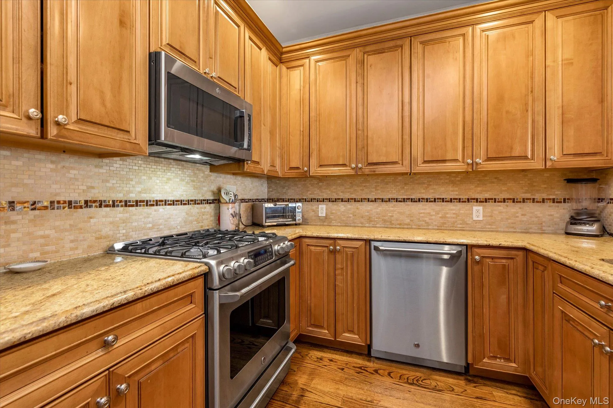 Kitchen featuring wood-finish cabinetry, light-toned countertops, and a tiled backsplash with a decorative accent strip Kitchen featuring wood-finish cabinetry, light-toned countertops, and a tiled backsplash with a decorative accent strip
