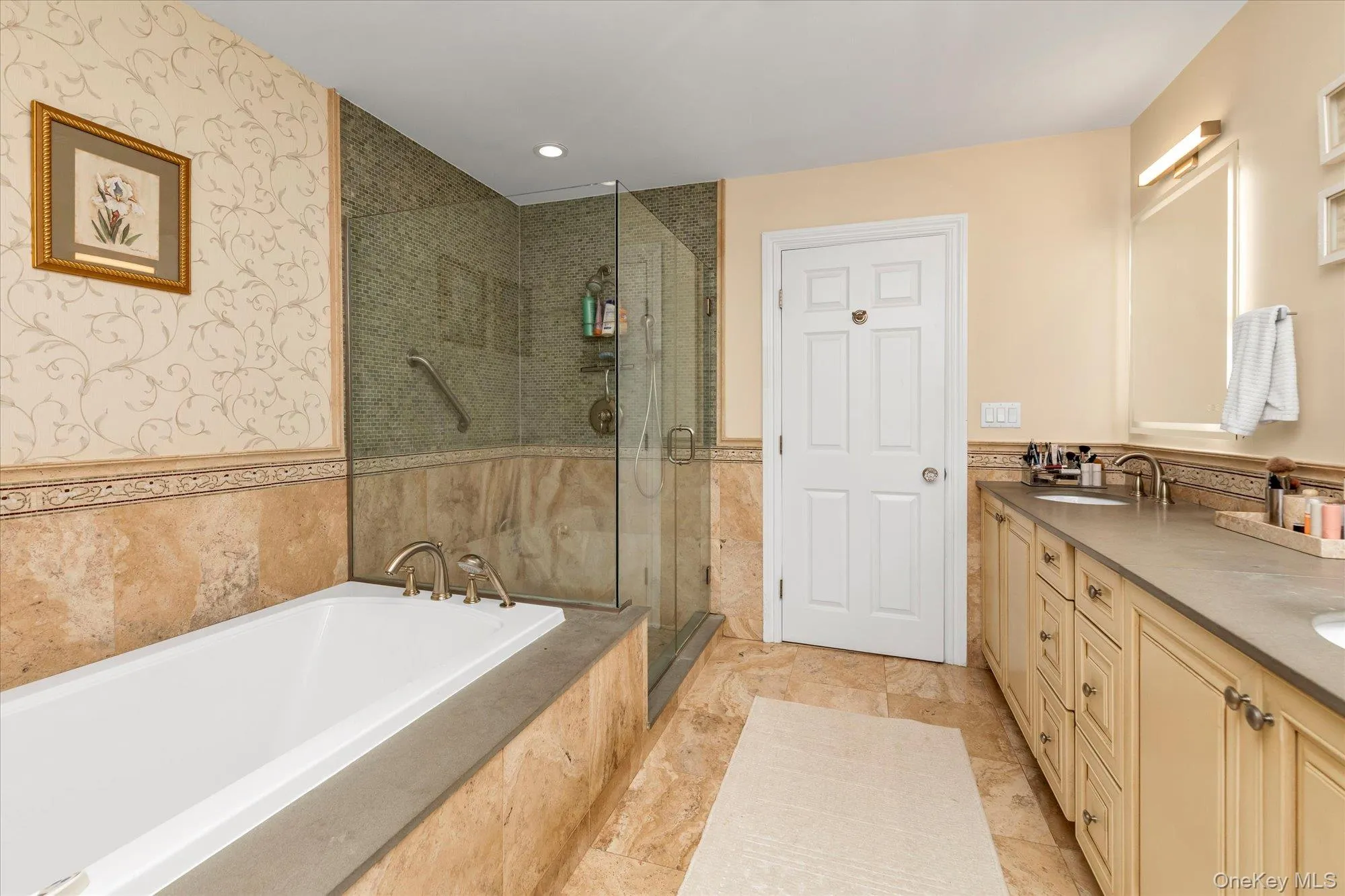 Bathroom featuring a built-in soaking tub, a glass-enclosed shower with mosaic tile, a dual vanity with undermount sinks, and stone-look tile flooring Bathroom featuring a built-in soaking tub, a glass-enclosed shower with mosaic tile, a dual vanity with undermount sinks, and stone-look tile flooring