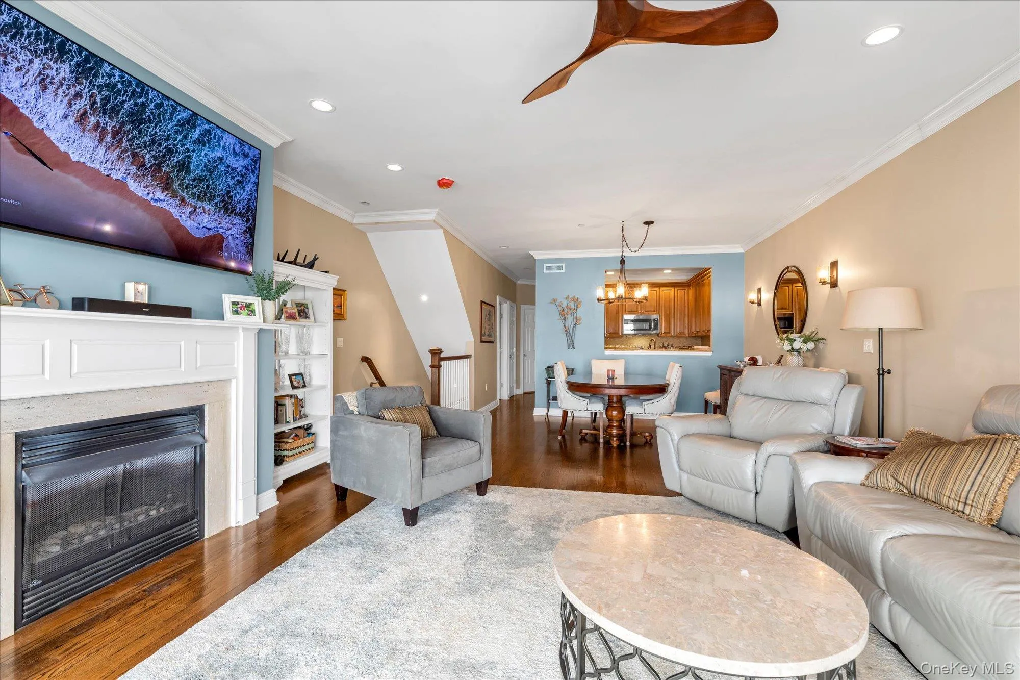 Living area featuring hardwood flooring, a fireplace with a white mantel, recessed lighting, and crown molding-view into the open dining area and kitchen Living area featuring hardwood flooring, a fireplace with a white mantel, recessed lighting, and crown molding-view into the open dining area and kitchen