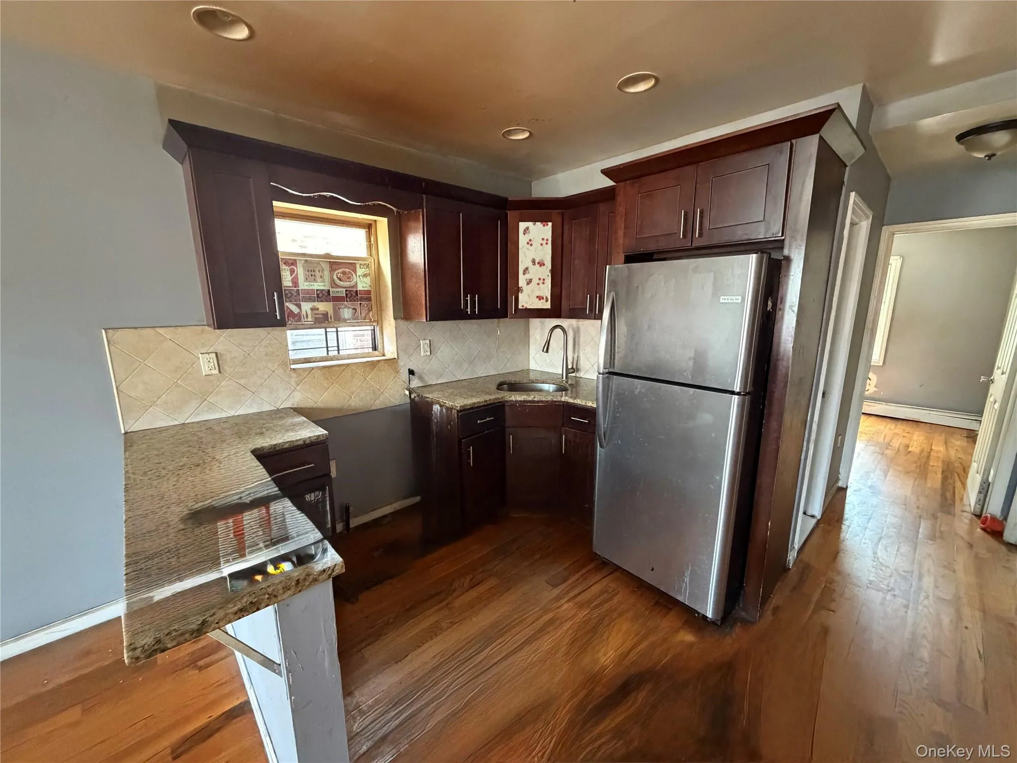 Kitchen featuring rich wood-finish cabinetry, granite-look countertops, a tiled backsplash, stainless steel refrigerator, and wood-finish flooring Kitchen featuring rich wood-finish cabinetry, granite-look countertops, a tiled backsplash, stainless steel refrigerator, and wood-finish flooring