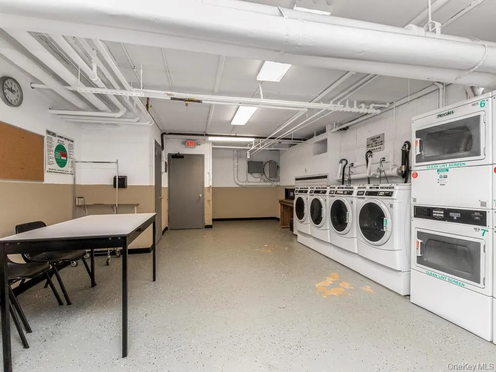 Dedicated laundry facility featuring multiple front-loading washing machines and stacked dryers, exposed overhead piping, concrete flooring, and a utility table Dedicated laundry facility featuring multiple front-loading washing machines and stacked dryers, exposed overhead piping, concrete flooring, and a utility table