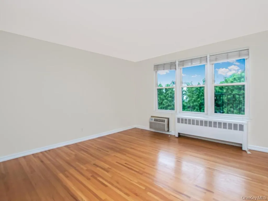 Room featuring expansive windows with white trim, wood-finish flooring, light neutral wall paint, white baseboards, and a baseboard heating unit Room featuring expansive windows with white trim, wood-finish flooring, light neutral wall paint, white baseboards, and a baseboard heating unit