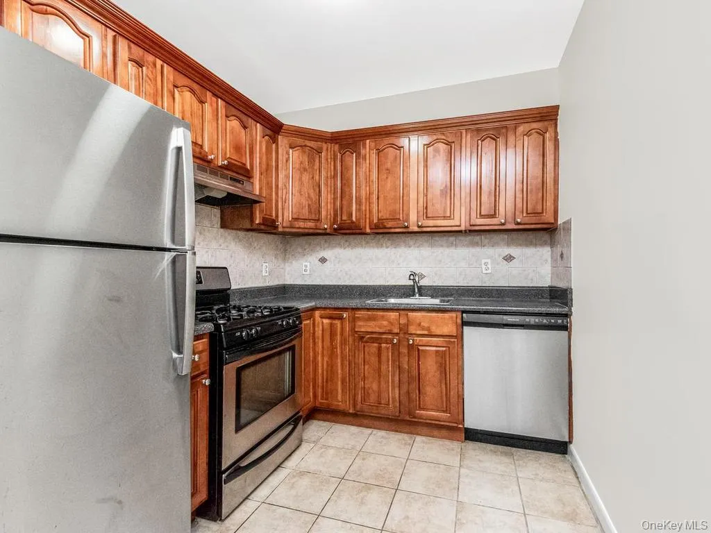 Kitchen featuring wood cabinetry, stainless steel appliances, dark countertops, and tiled flooring Kitchen featuring wood cabinetry, stainless steel appliances, dark countertops, and tiled flooring