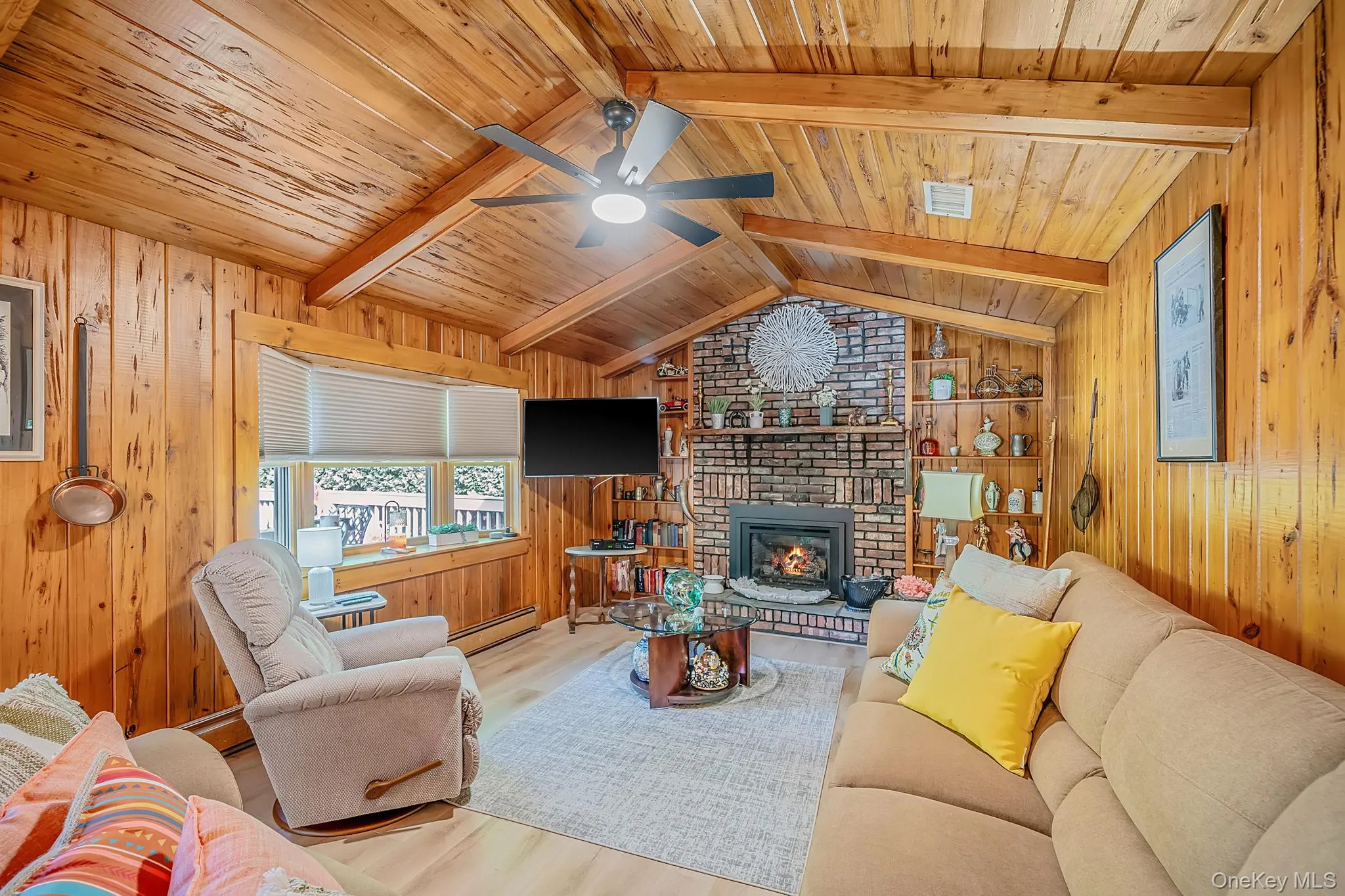 Vaulted wood-paneled ceiling with exposed beams, brick fireplace with a hearth, window with horizontal blinds, and wood-finish flooring Vaulted wood-paneled ceiling with exposed beams, brick fireplace with a hearth, window with horizontal blinds, and wood-finish flooring
