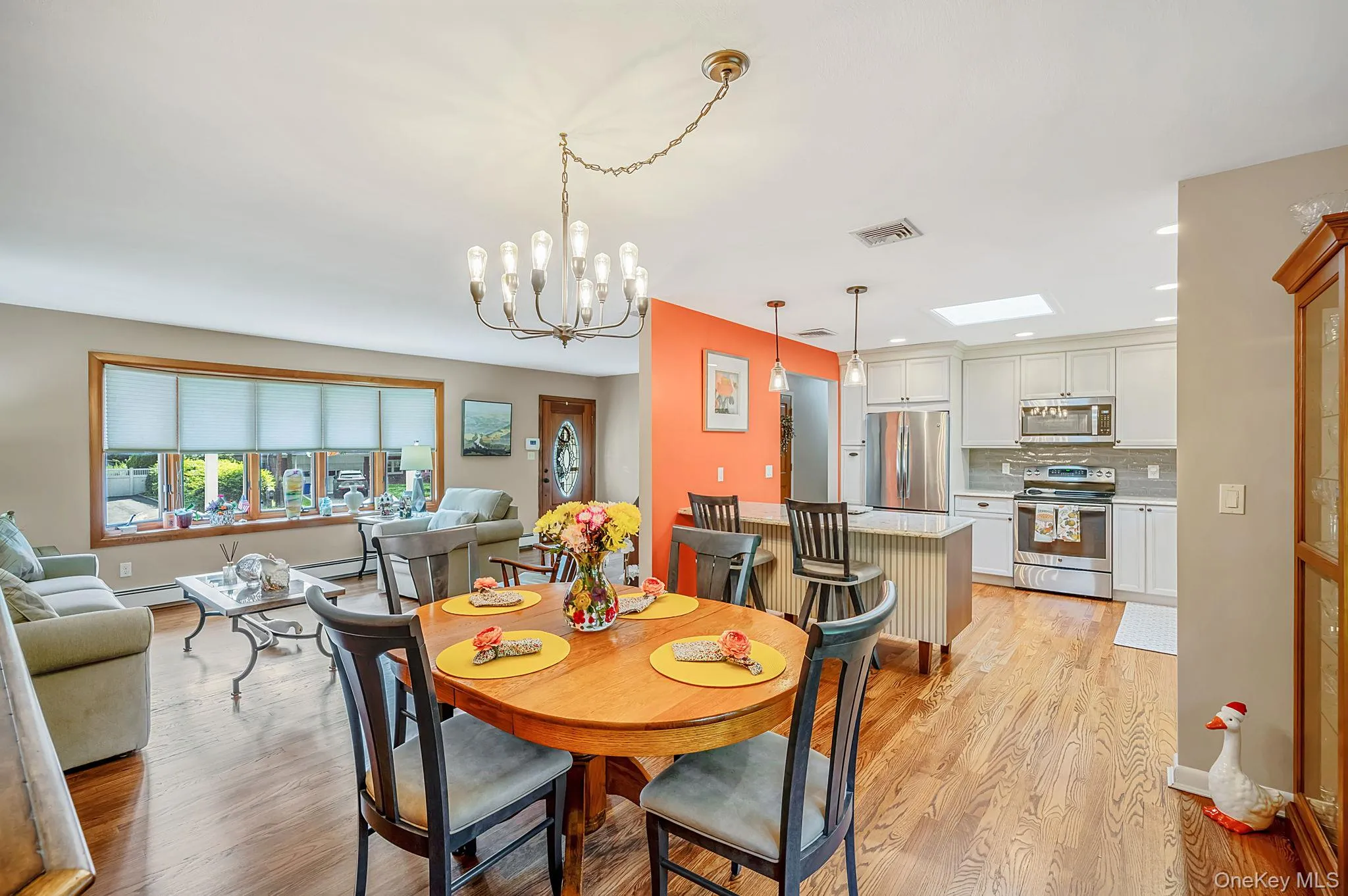 Open-concept living and dining area featuring hardwood flooring, a wood-finish oval dining table with seating for four, an elegant chandelier with candle-style lights, and abundant natural light from large multi-pane windows Open-concept living and dining area featuring hardwood flooring, a wood-finish oval dining table with seating for four, an elegant chandelier with candle-style lights, and abundant natural light from large multi-pane windows
