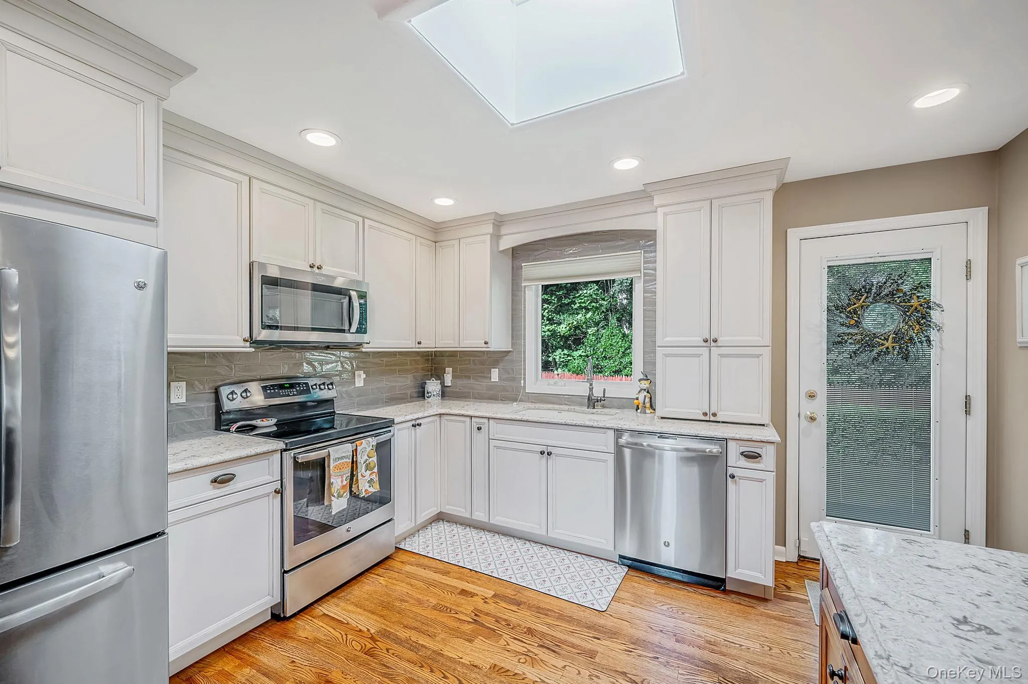 Kitchen featuring white cabinetry, granite countertops, stainless steel appliances, wood-finish flooring, and a skylight Kitchen featuring white cabinetry, granite countertops, stainless steel appliances, wood-finish flooring, and a skylight