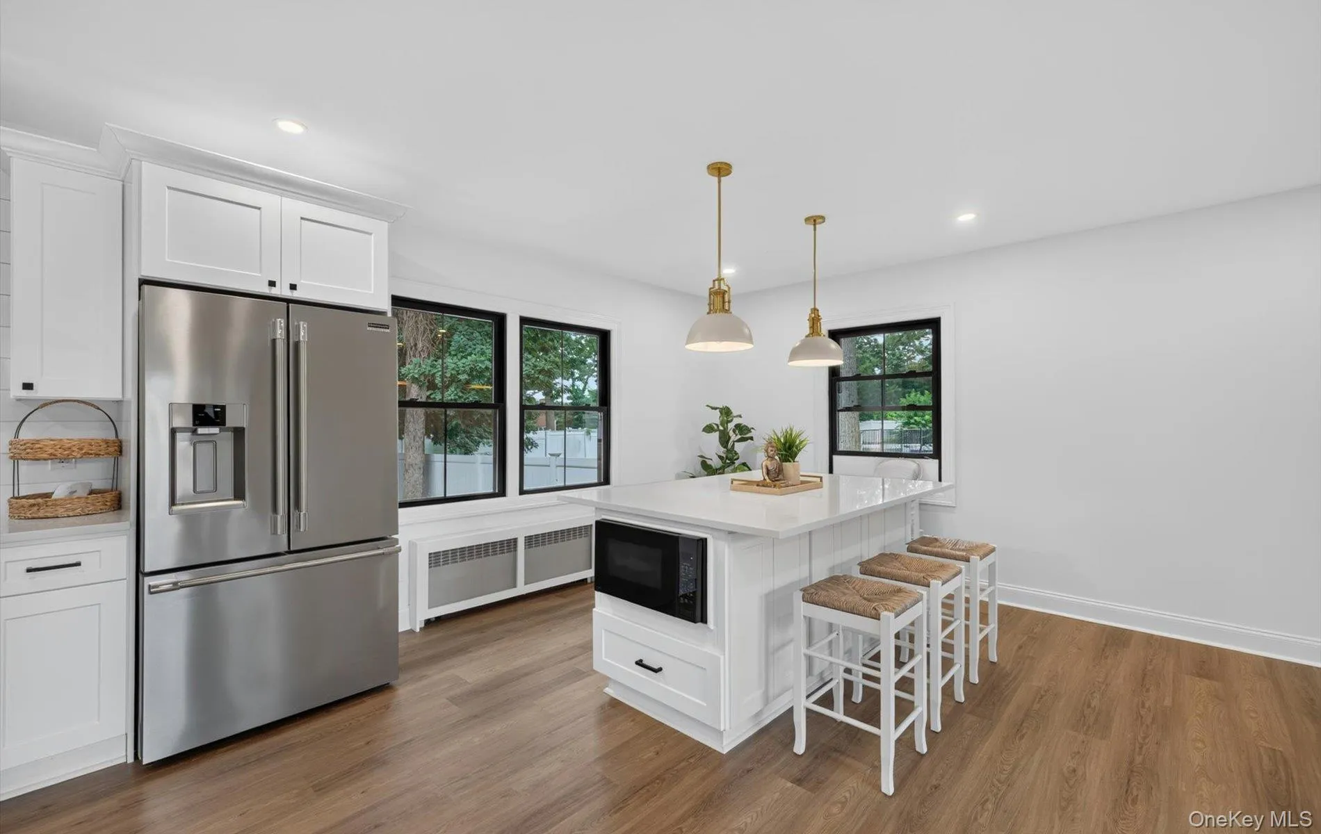 Modern kitchen featuring a central island with integrated microwave, white shaker-style cabinetry, stainless steel refrigerator, wood-finish flooring, and pendant lighting Modern kitchen featuring a central island with integrated microwave, white shaker-style cabinetry, stainless steel refrigerator, wood-finish flooring, and pendant lighting