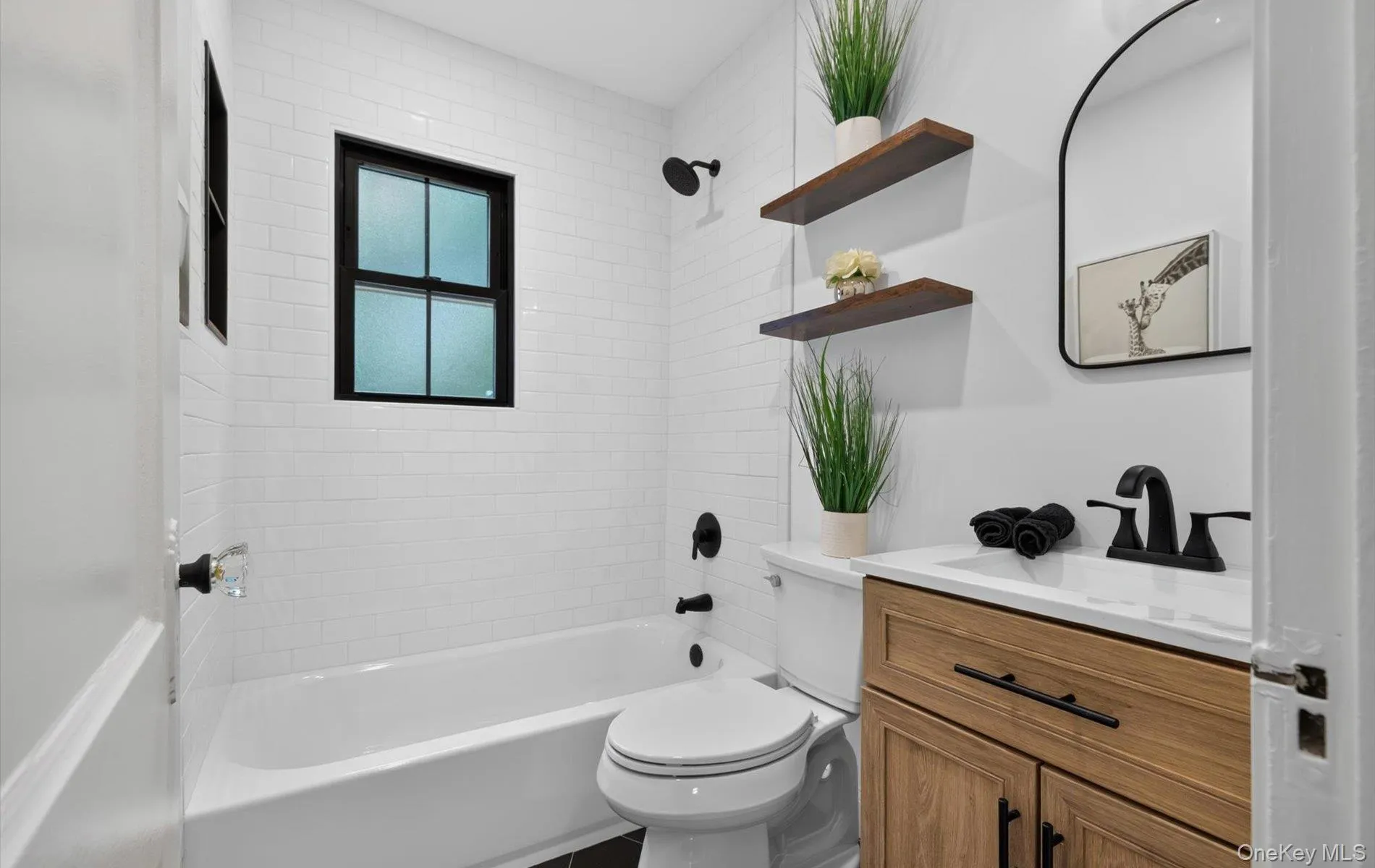 Full bathroom with white subway tile walls, a single window with dark framing, a built-in tub with dark fixtures, a wooden vanity with a white countertop, and a black-framed wall mirror Full bathroom with white subway tile walls, a single window with dark framing, a built-in tub with dark fixtures, a wooden vanity with a white countertop, and a black-framed wall mirror