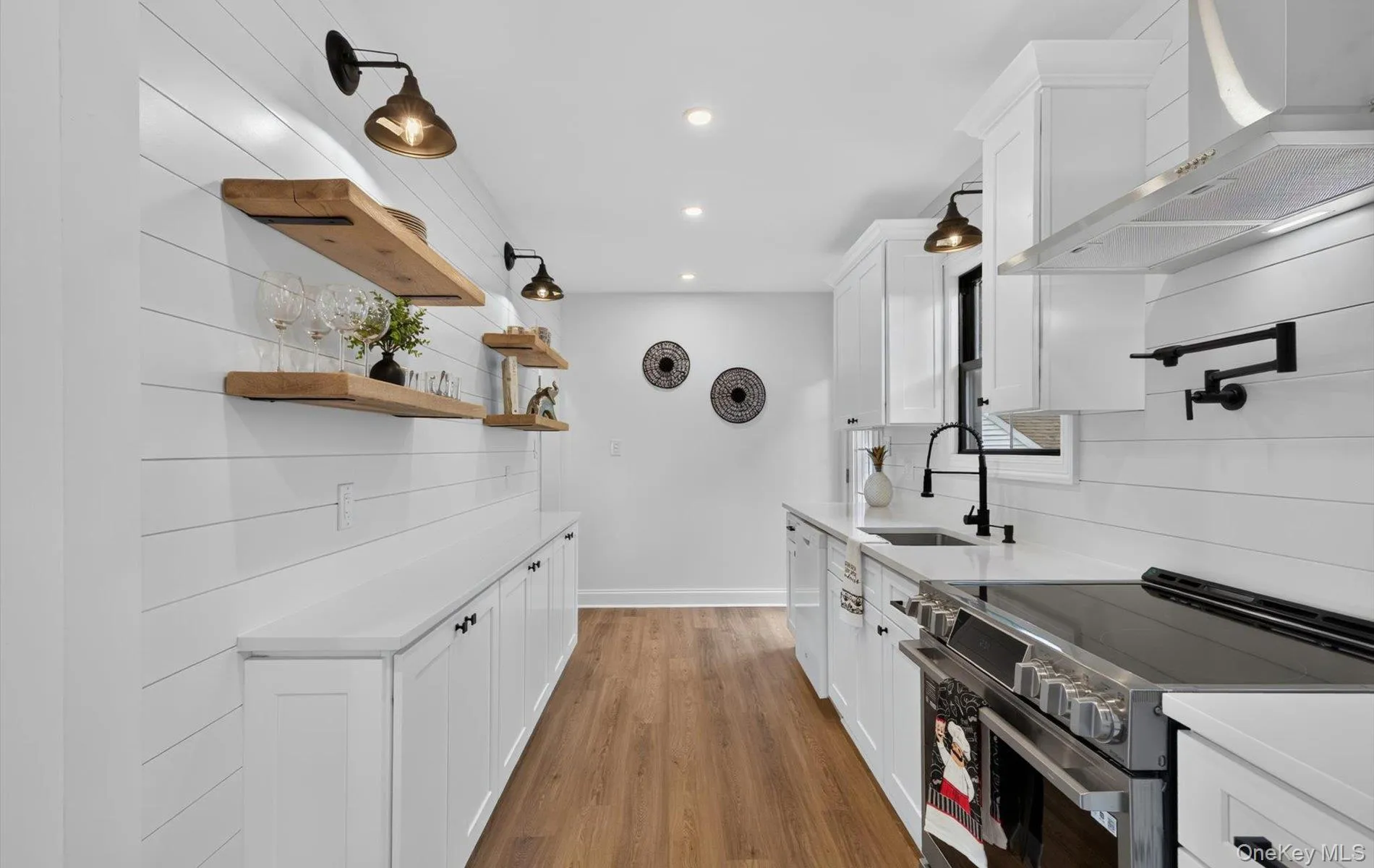 Modern kitchen featuring white shaker-style cabinetry, a stainless steel range with a pot filler, a matching range hood, wood-finish flooring, and shiplap wall finishes Modern kitchen featuring white shaker-style cabinetry, a stainless steel range with a pot filler, a matching range hood, wood-finish flooring, and shiplap wall finishes