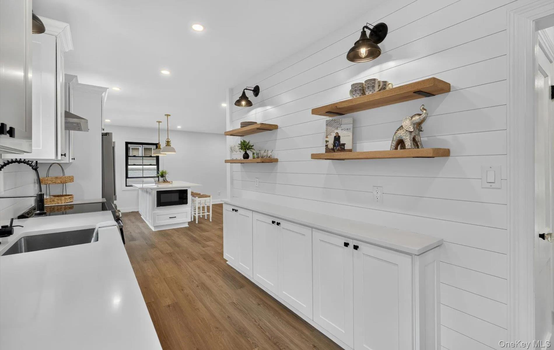 Spacious kitchen featuring white cabinetry, light countertops, wood-finish flooring, a central island with built-in microwave, and open wood shelving on a shiplap accent wall Spacious kitchen featuring white cabinetry, light countertops, wood-finish flooring, a central island with built-in microwave, and open wood shelving on a shiplap accent wall