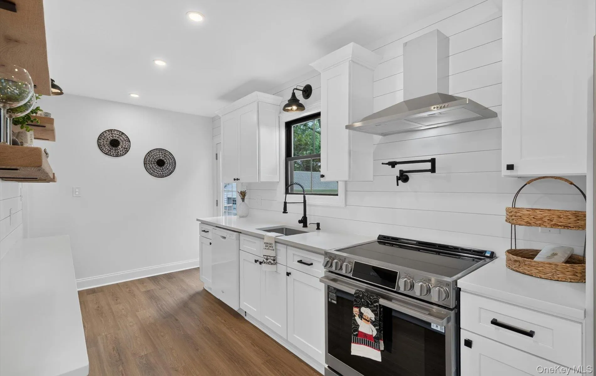 Modern kitchen featuring white shaker cabinetry, stainless steel range with coordinating range hood, white shiplap backsplash, wood-finish flooring, and recessed lighting Modern kitchen featuring white shaker cabinetry, stainless steel range with coordinating range hood, white shiplap backsplash, wood-finish flooring, and recessed lighting