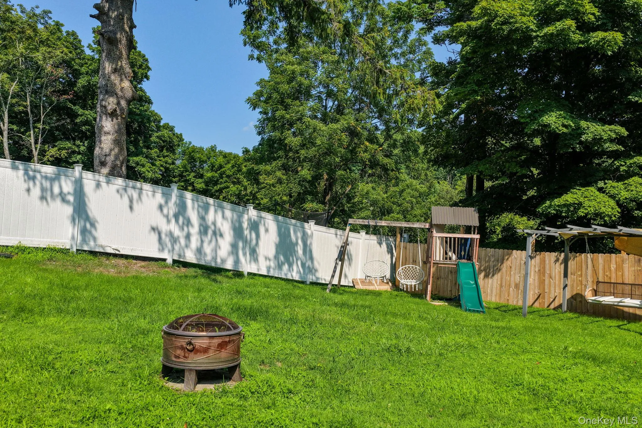 Expansive lawn area featuring a white privacy fence, mature trees, and a fire pit Expansive lawn area featuring a white privacy fence, mature trees, and a fire pit