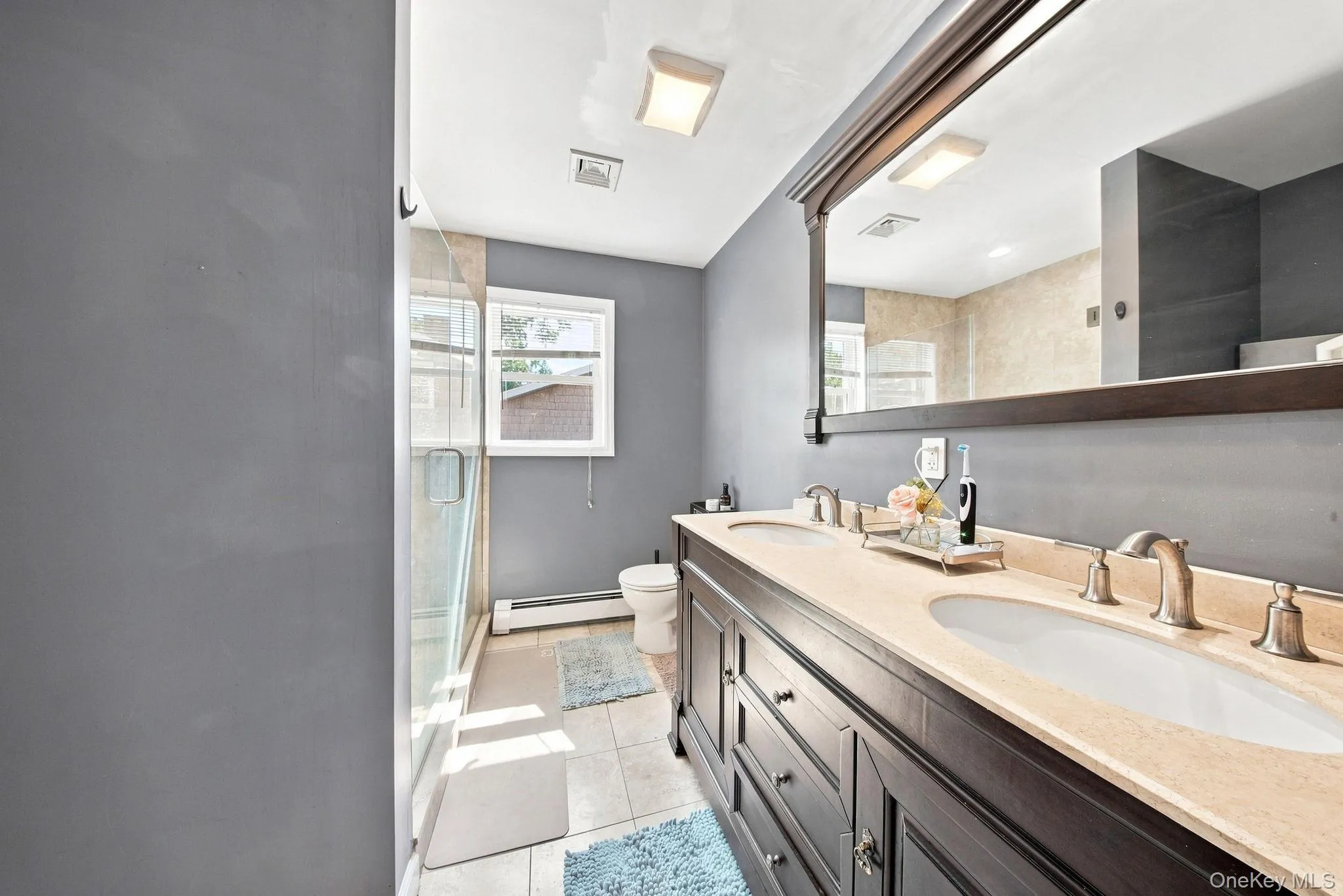 Spa-inspired primary bathroom featuring dual vanities with wood-finish cabinetry, cream-colored countertops, and brushed nickel fixtures Spa-inspired primary bathroom featuring dual vanities with wood-finish cabinetry, cream-colored countertops, and brushed nickel fixtures