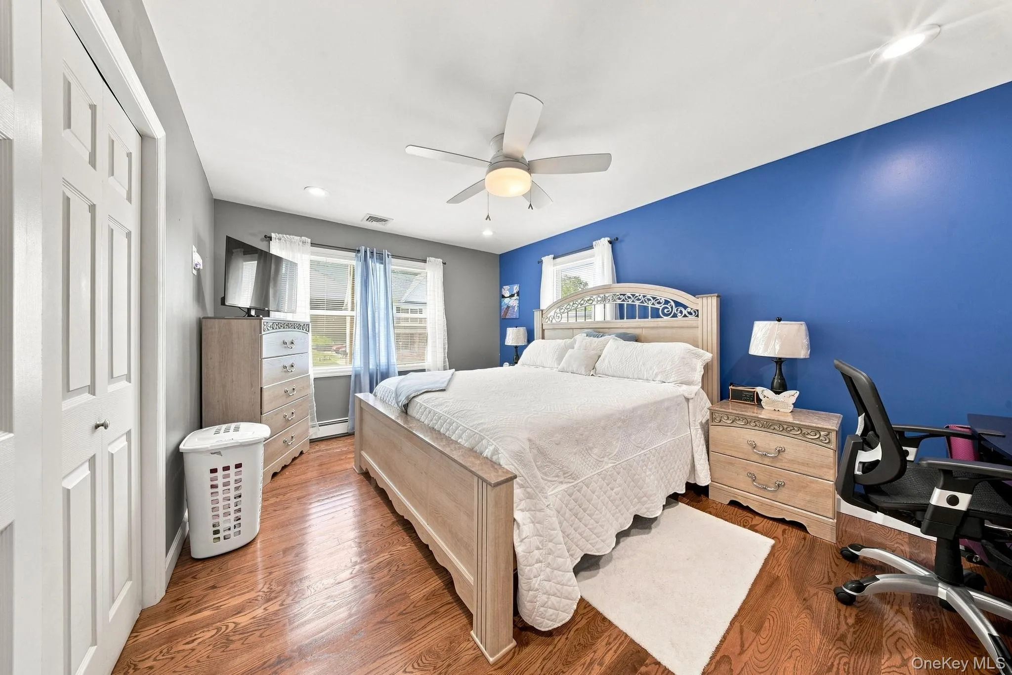 Spacious bedroom featuring rich wood-finish flooring, warm wood-tone bed frame and nightstands, and a striking cobalt blue accent wall Spacious bedroom featuring rich wood-finish flooring, warm wood-tone bed frame and nightstands, and a striking cobalt blue accent wall