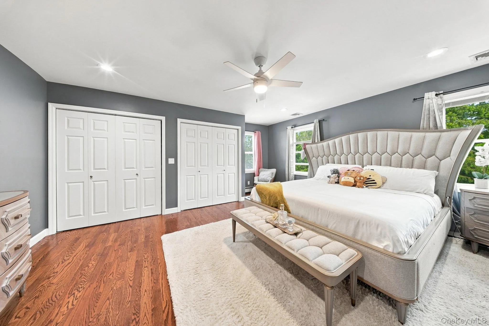 Spacious primary bedroom featuring a tufted upholstered headboard, hardwood flooring, dual walk-in closets with six-panel doors, ceiling fan with integrated lighting, and abundant natural light from multiple windows overlooking the landscape Spacious primary bedroom featuring a tufted upholstered headboard, hardwood flooring, dual walk-in closets with six-panel doors, ceiling fan with integrated lighting, and abundant natural light from multiple windows overlooking the landscape