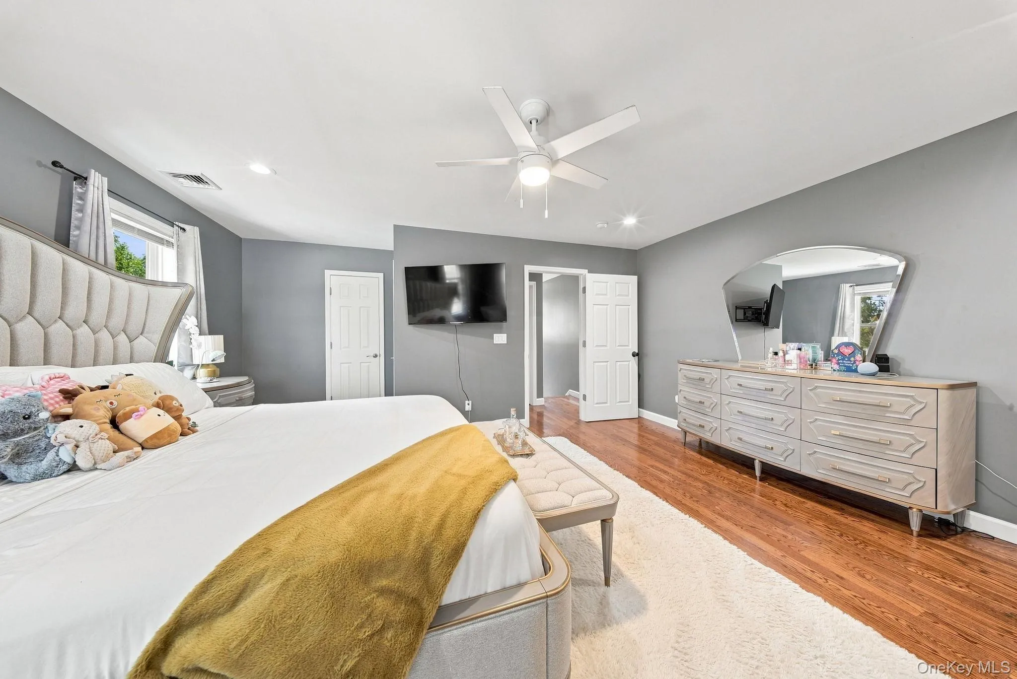 Spacious primary bedroom with gray walls, white five-panel doors, wood-finish flooring, and ceiling fan with integrated lighting Spacious primary bedroom with gray walls, white five-panel doors, wood-finish flooring, and ceiling fan with integrated lighting