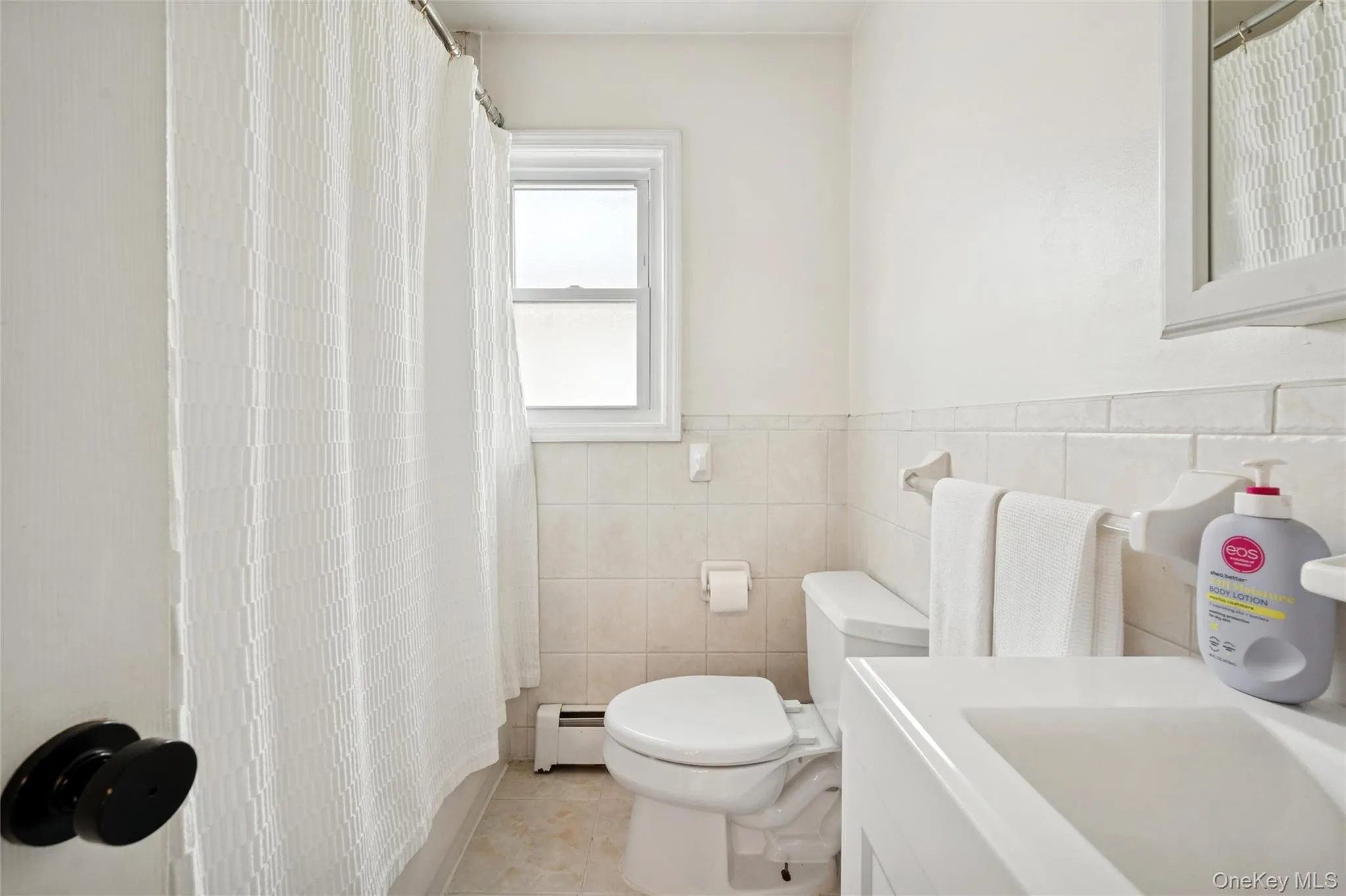 Full bath featuring a shower with shower curtain, vanity, a baseboard radiator, light tile patterned floors, and tile walls Full bath featuring a shower with shower curtain, vanity, a baseboard radiator, light tile patterned floors, and tile walls