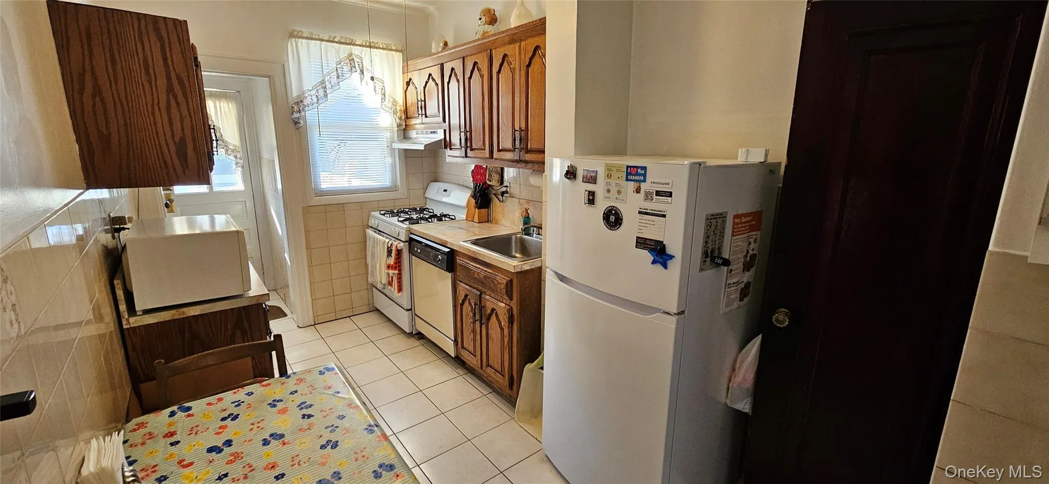 Kitchen with white appliances, light tile patterned floors, light countertops, brown cabinetry, and under cabinet range hood Kitchen with white appliances, light tile patterned floors, light countertops, brown cabinetry, and under cabinet range hood