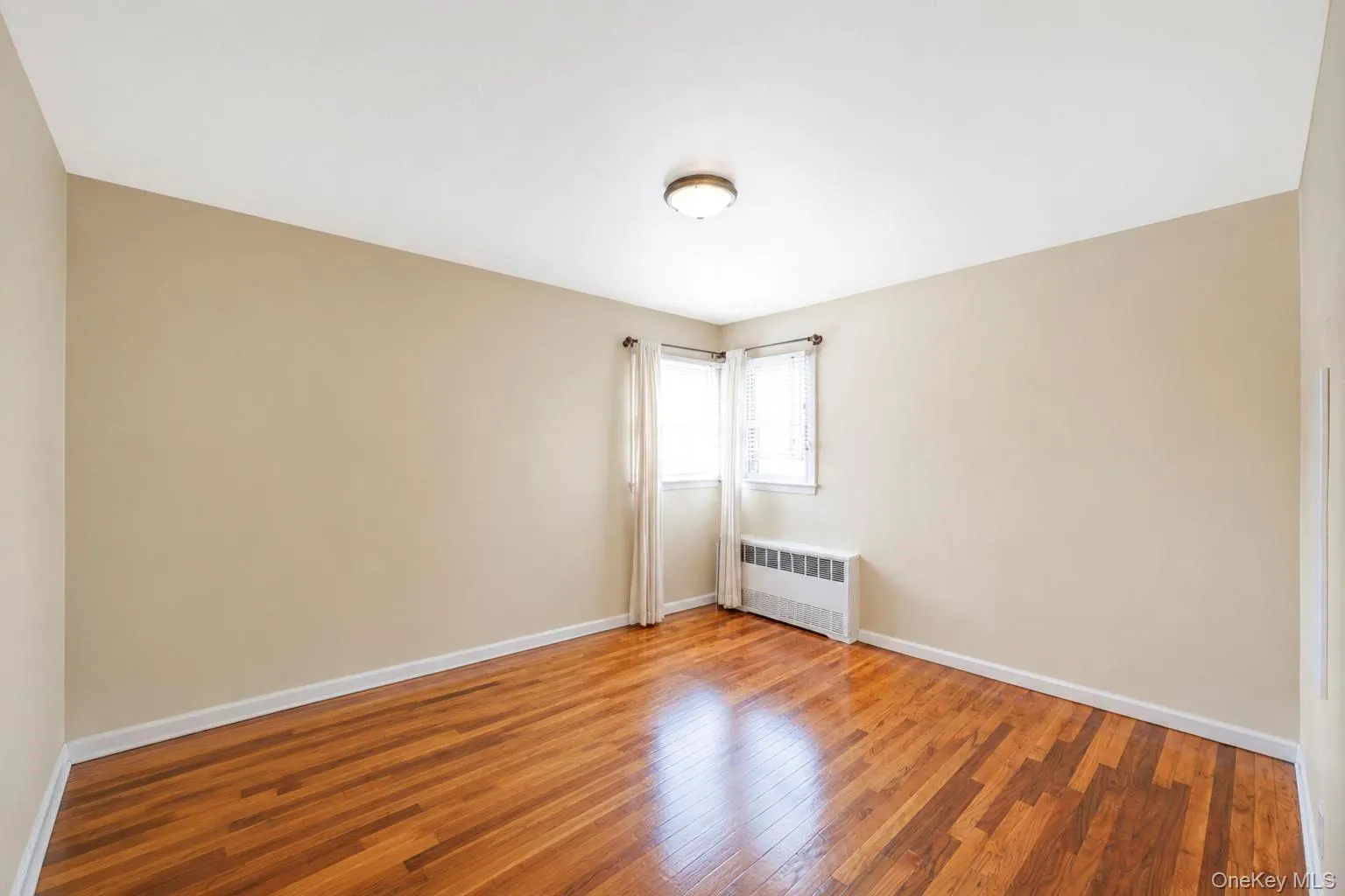 Room featuring wood-finish flooring, neutral wall paint, white trim, two windows, and a ceiling-mounted light fixture Room featuring wood-finish flooring, neutral wall paint, white trim, two windows, and a ceiling-mounted light fixture