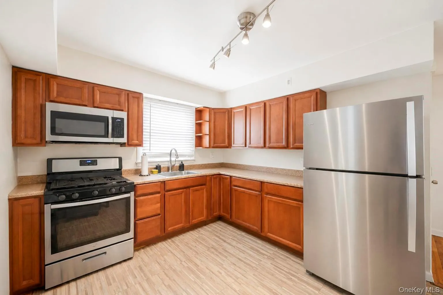 Kitchen featuring wood cabinetry, stainless steel appliances, track lighting, light-toned countertops, and wood-finish flooring Kitchen featuring wood cabinetry, stainless steel appliances, track lighting, light-toned countertops, and wood-finish flooring