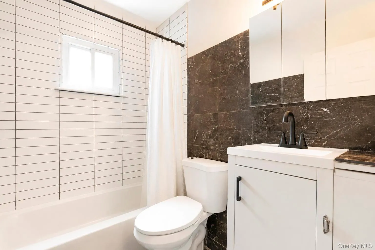 Bathroom featuring dark marble-look wall tiling, white vanity with matte black hardware, dual mirrored medicine cabinets, and a white subway-tiled shower with a window Bathroom featuring dark marble-look wall tiling, white vanity with matte black hardware, dual mirrored medicine cabinets, and a white subway-tiled shower with a window