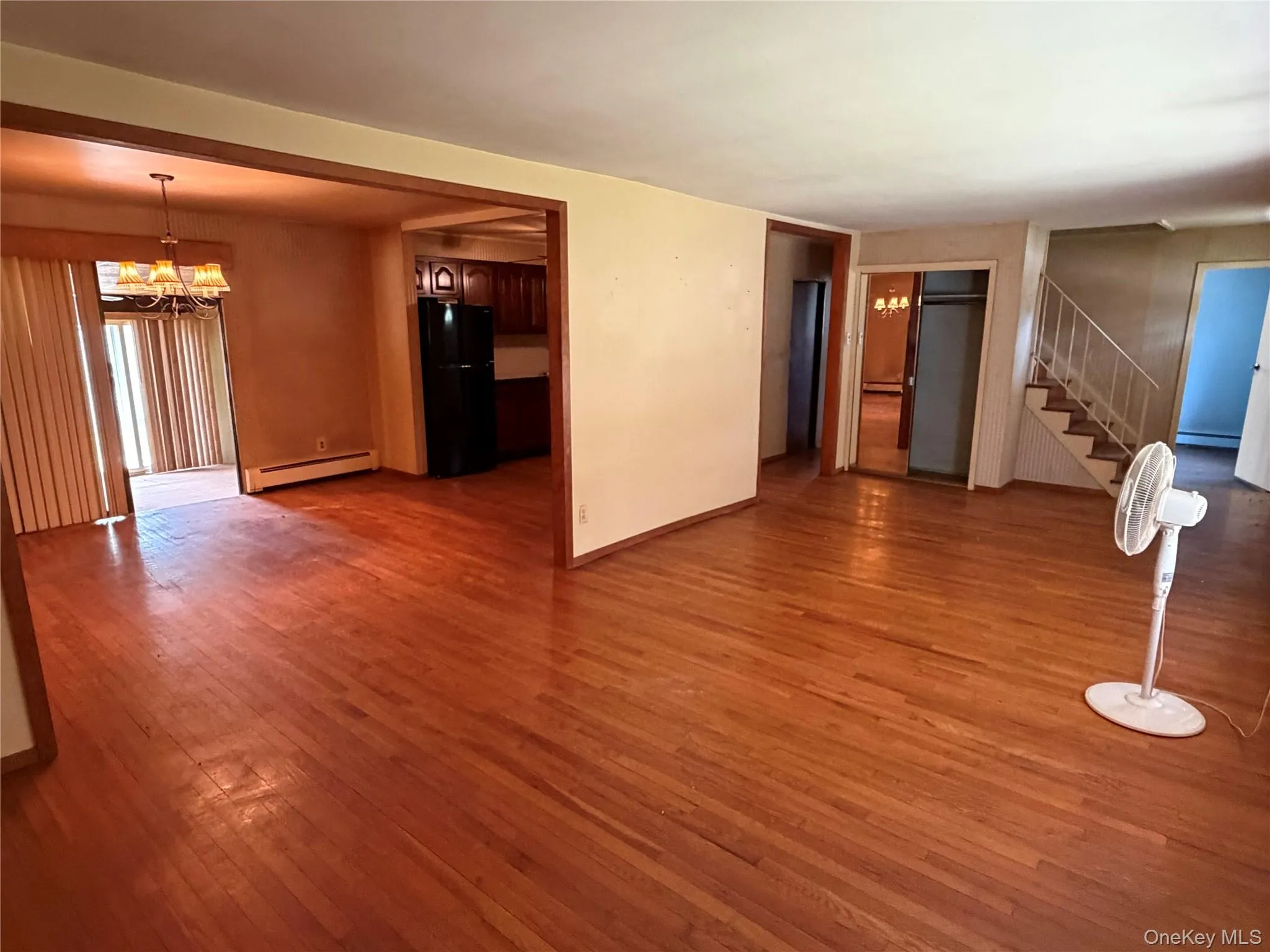 Expansive main living area featuring wood-finish flooring, a staircase with metal railings, and a doorway leading to a kitchen with dark cabinetry and a black refrigerator Expansive main living area featuring wood-finish flooring, a staircase with metal railings, and a doorway leading to a kitchen with dark cabinetry and a black refrigerator