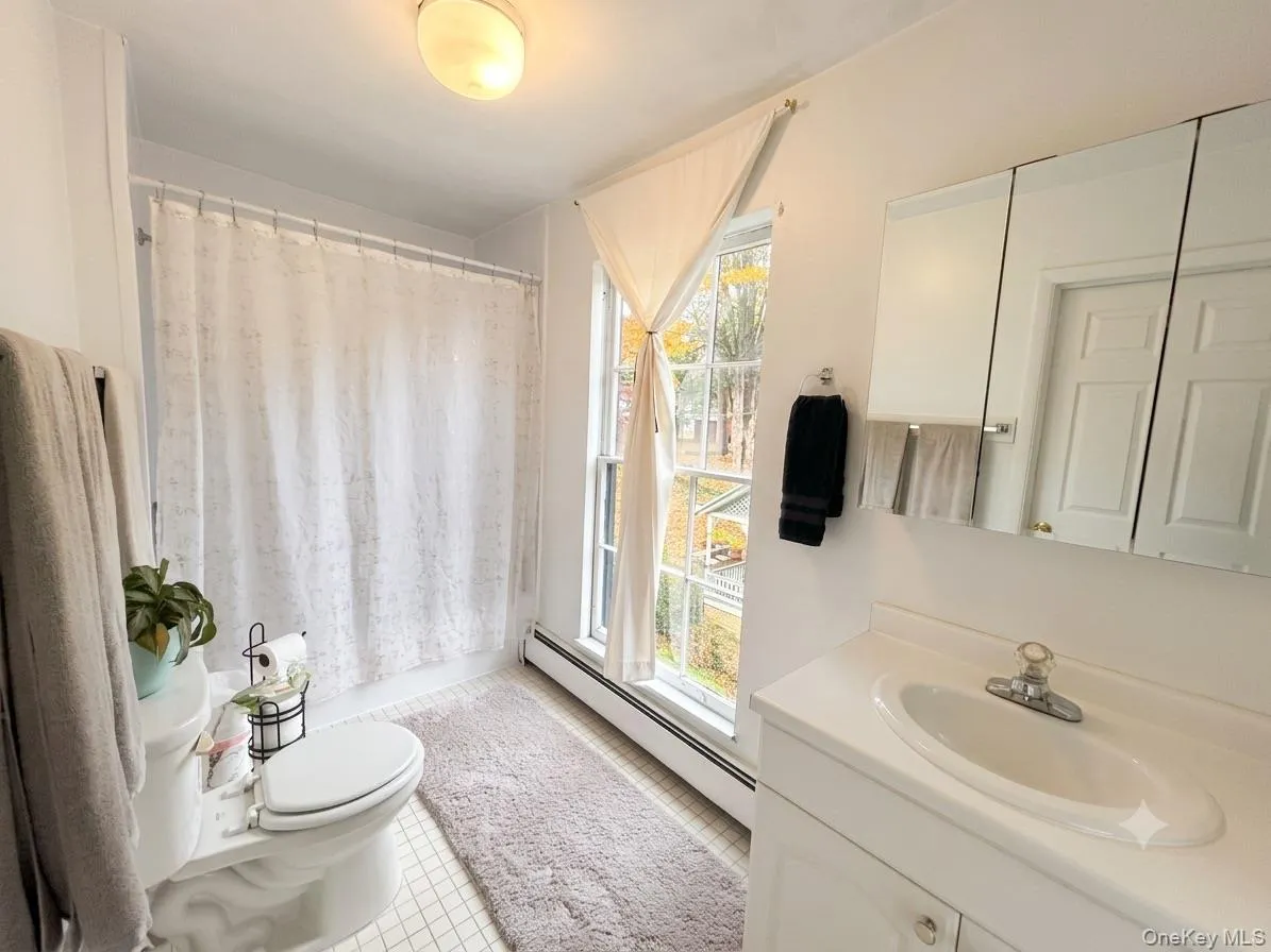 Bathroom featuring a white vanity with an integrated sink, a mirrored medicine cabinet, a full bathtub with shower, and white square tile flooring Bathroom featuring a white vanity with an integrated sink, a mirrored medicine cabinet, a full bathtub with shower, and white square tile flooring
