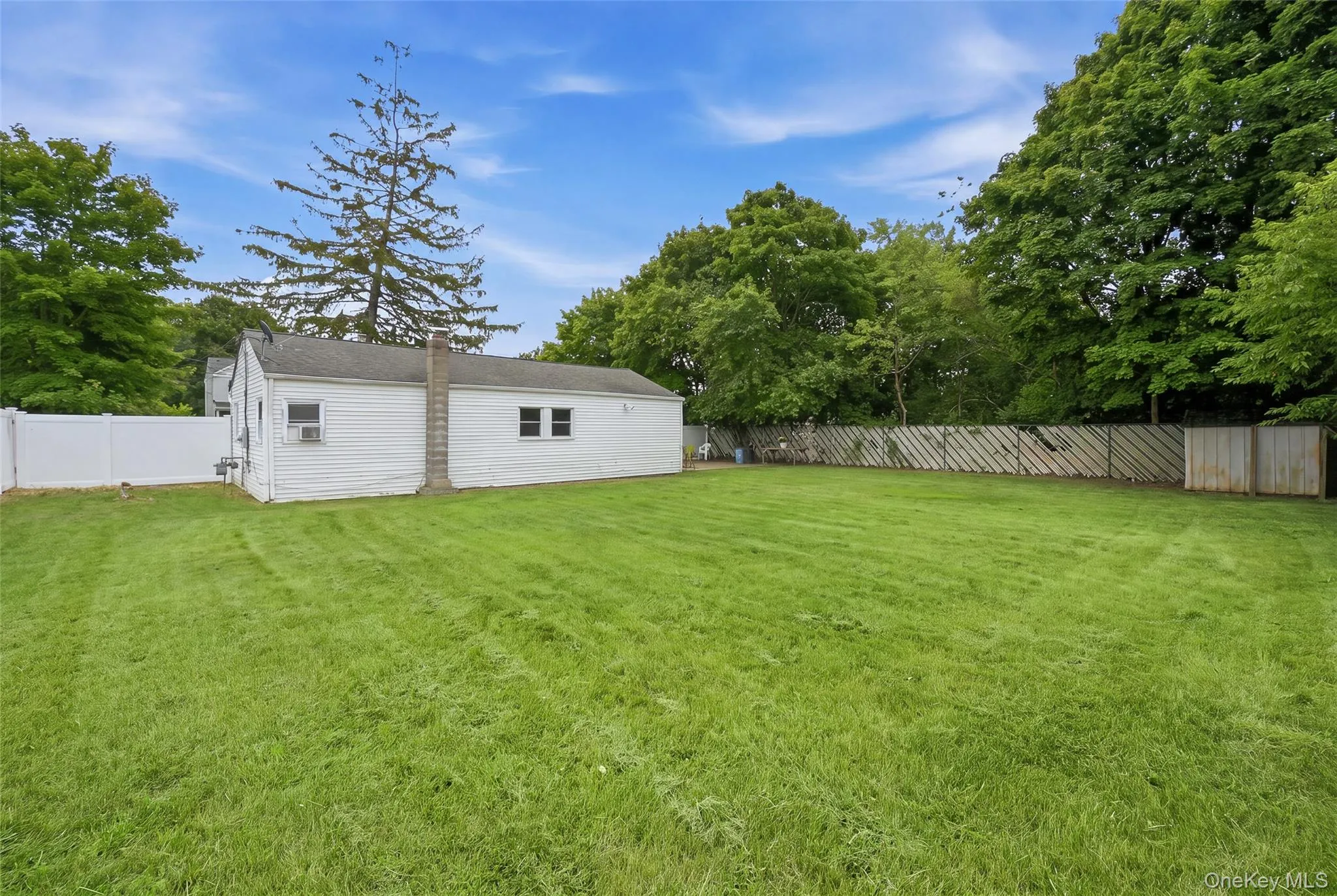 Spacious backyard featuring a large green lawn, partial white vinyl fencing, and a side view of the white siding exterior with a visible chimney Spacious backyard featuring a large green lawn, partial white vinyl fencing, and a side view of the white siding exterior with a visible chimney