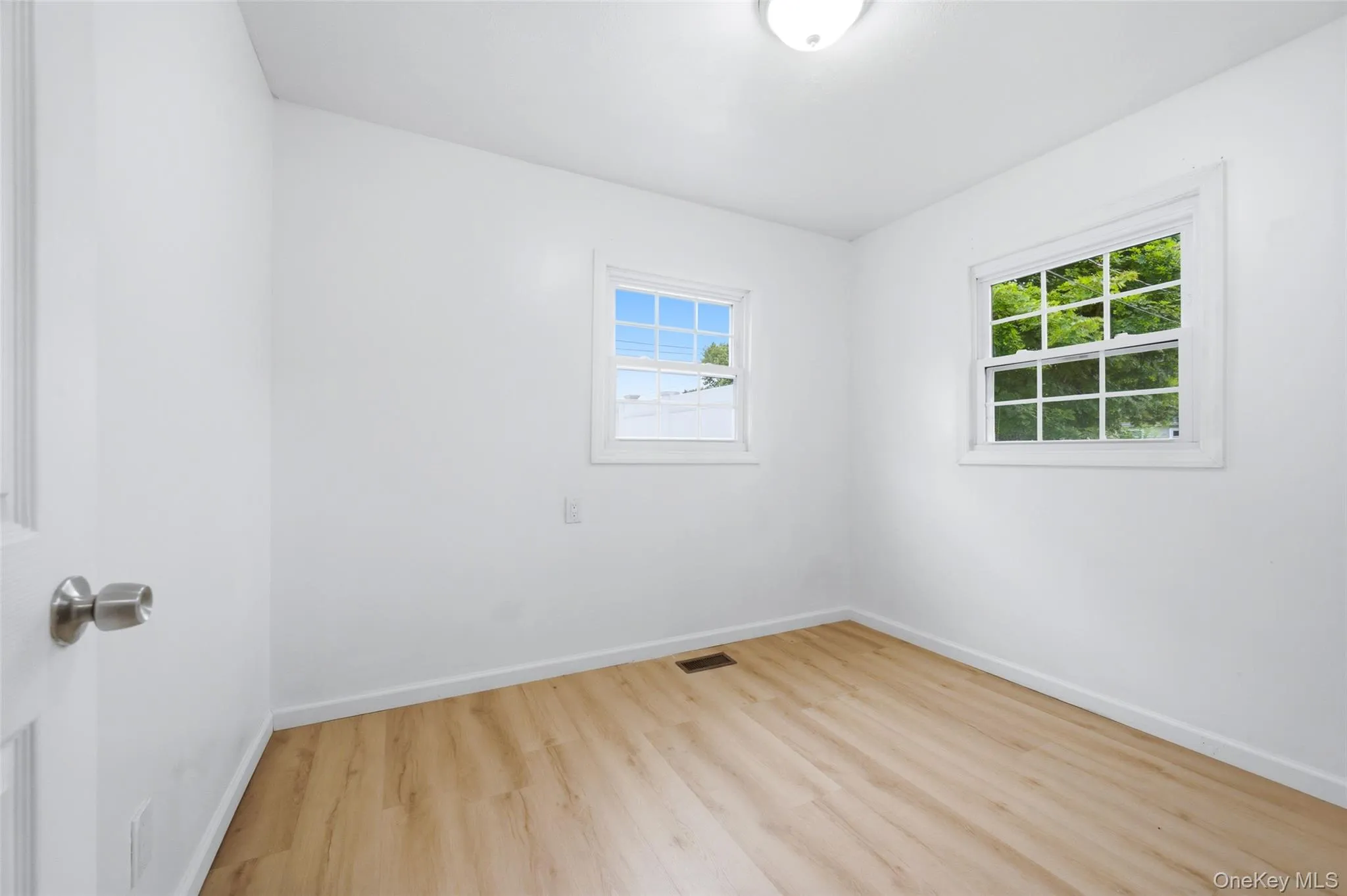 Bright room featuring wood-finish flooring, white walls, two double-hung windows, and a flush mount ceiling light fixture Bright room featuring wood-finish flooring, white walls, two double-hung windows, and a flush mount ceiling light fixture