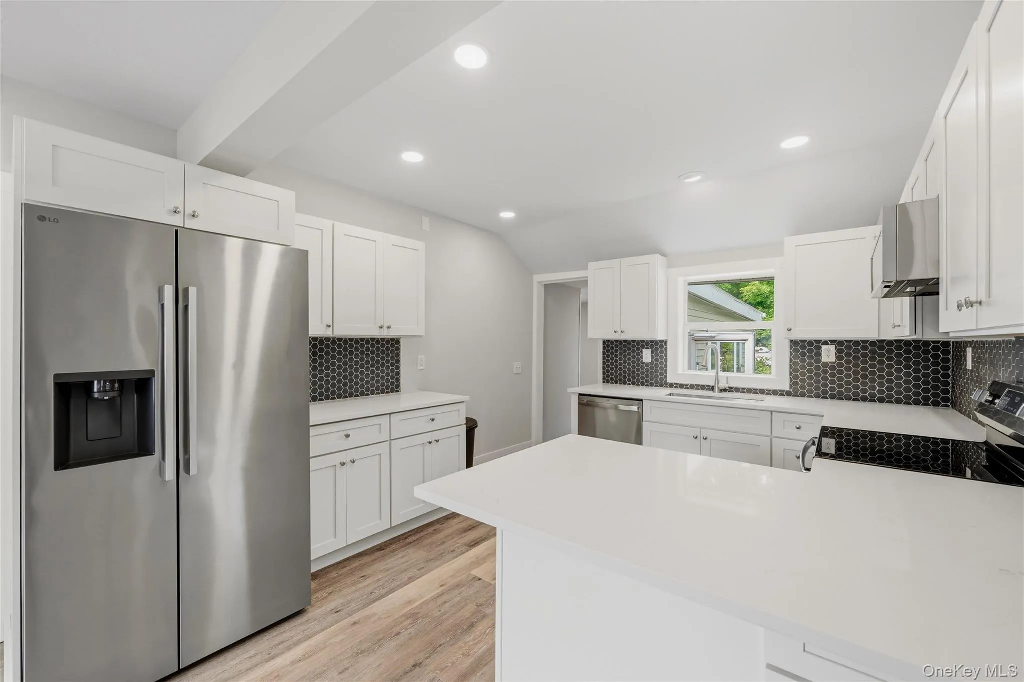 Modern kitchen featuring white cabinetry, a large white island with a solid surface countertop, stainless steel appliances, a dark hexagonal tile backsplash, and light wood-finish flooring Modern kitchen featuring white cabinetry, a large white island with a solid surface countertop, stainless steel appliances, a dark hexagonal tile backsplash, and light wood-finish flooring