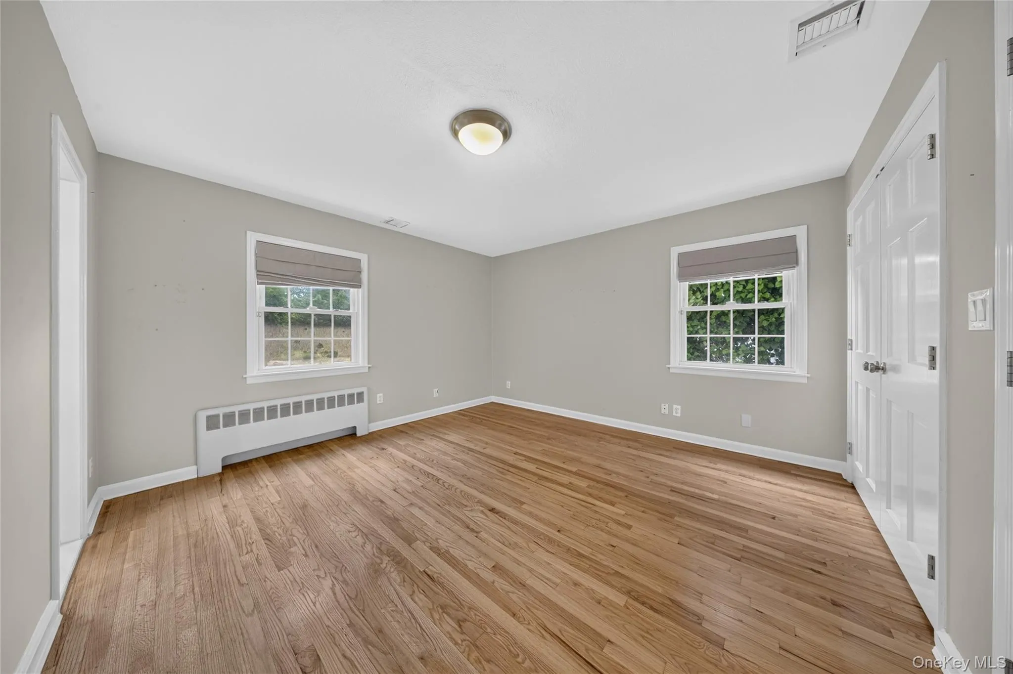 Hardwood flooring throughout this room featuring two windows with roman shades, a built-in radiator, and a flush mount ceiling light Hardwood flooring throughout this room featuring two windows with roman shades, a built-in radiator, and a flush mount ceiling light