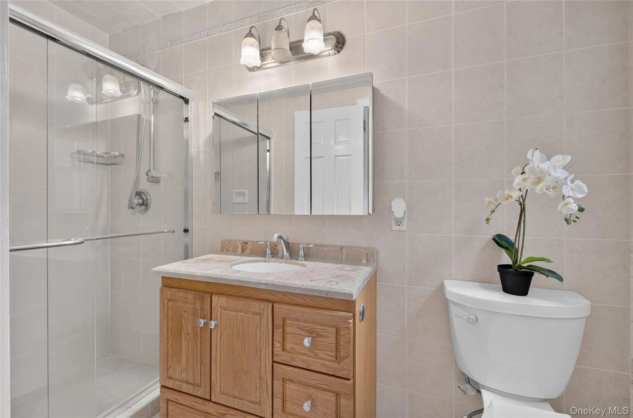 Full bathroom featuring a single-sink vanity with wood-finish cabinetry, a framed mirror, a three-light fixture, a walk-in shower with a glass enclosure, and tiled walls Full bathroom featuring a single-sink vanity with wood-finish cabinetry, a framed mirror, a three-light fixture, a walk-in shower with a glass enclosure, and tiled walls