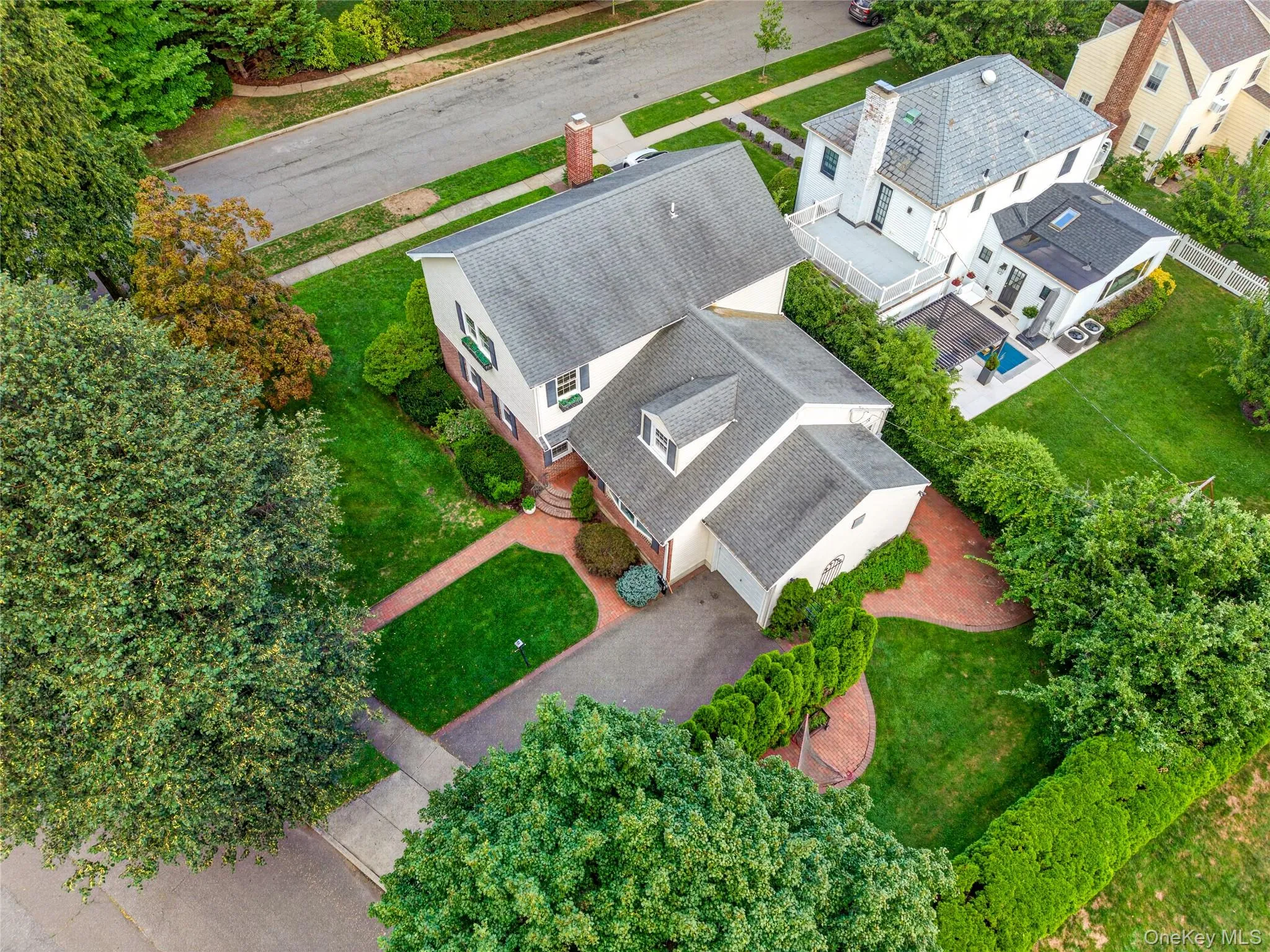 Traditional residence featuring a brick paver driveway, verdant landscaping, mature trees, and a manicured lawn Traditional residence featuring a brick paver driveway, verdant landscaping, mature trees, and a manicured lawn