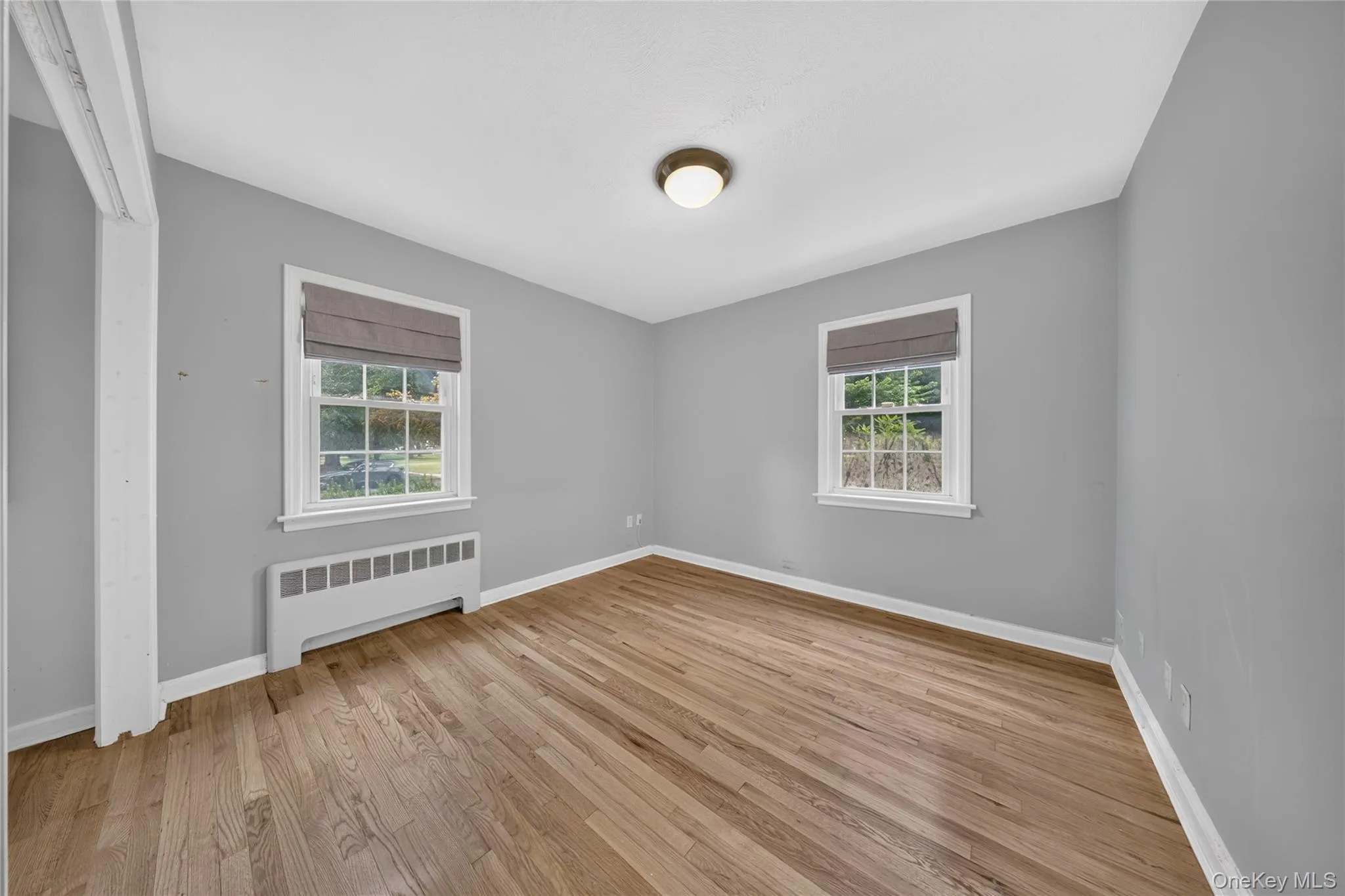Room featuring wood-finish flooring, two windows with blinds, a ceiling-mounted light fixture, light gray walls, and white baseboards Room featuring wood-finish flooring, two windows with blinds, a ceiling-mounted light fixture, light gray walls, and white baseboards