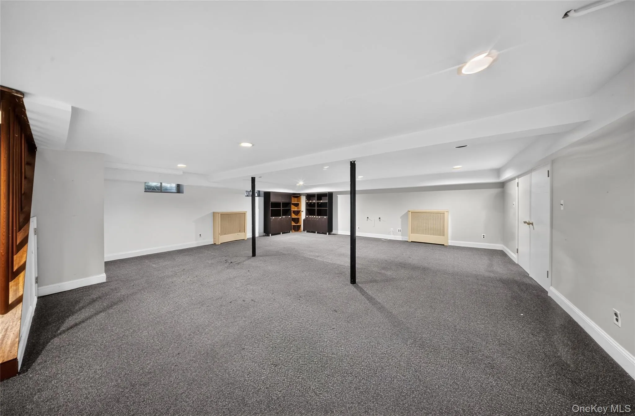 Spacious finished basement featuring dark gray carpeting, recessed lighting, built-in shelving, two white panel doors, and a small egress window Spacious finished basement featuring dark gray carpeting, recessed lighting, built-in shelving, two white panel doors, and a small egress window