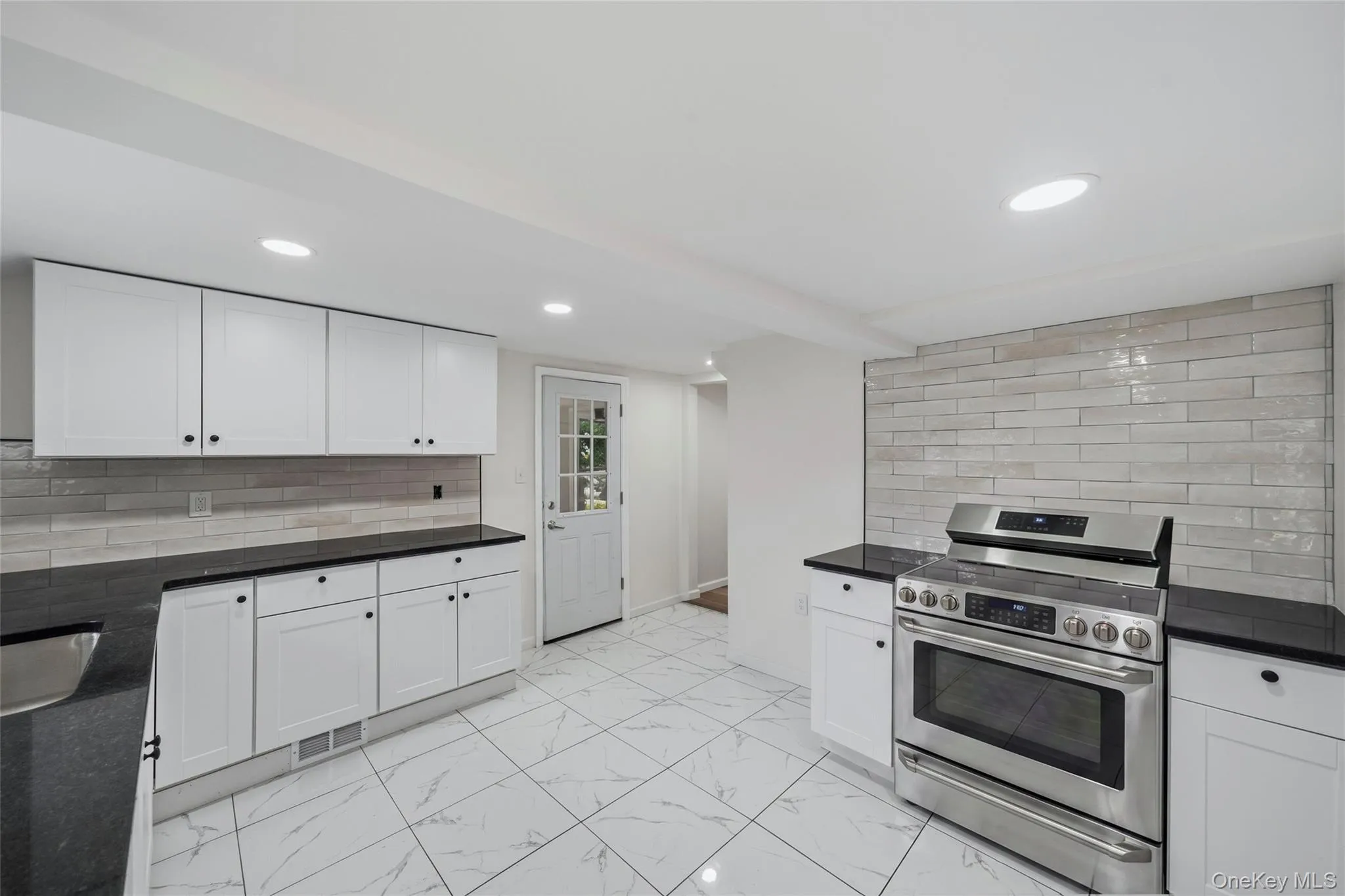 Modern kitchen featuring white cabinetry with black hardware and countertops, stainless steel gas range with convection oven, ceramic tile flooring in a neutral palette, recessed ceiling lighting, and subway tile backsplash Modern kitchen featuring white cabinetry with black hardware and countertops, stainless steel gas range with convection oven, ceramic tile flooring in a neutral palette, recessed ceiling lighting, and subway tile backsplash