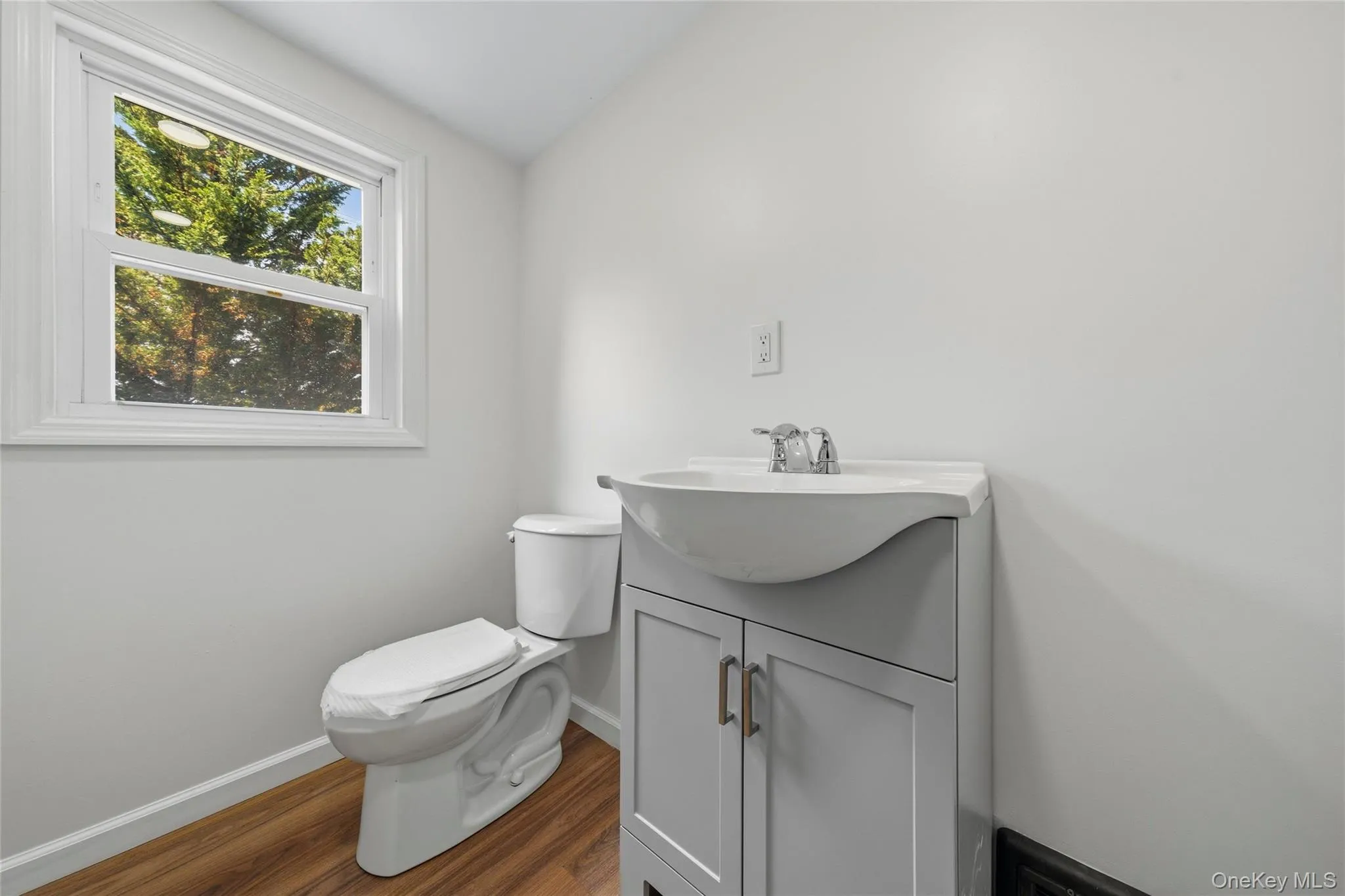 Bathroom featuring a window, wood-finish flooring, a white toilet, and a vanity with a white basin and grey cabinetry Bathroom featuring a window, wood-finish flooring, a white toilet, and a vanity with a white basin and grey cabinetry