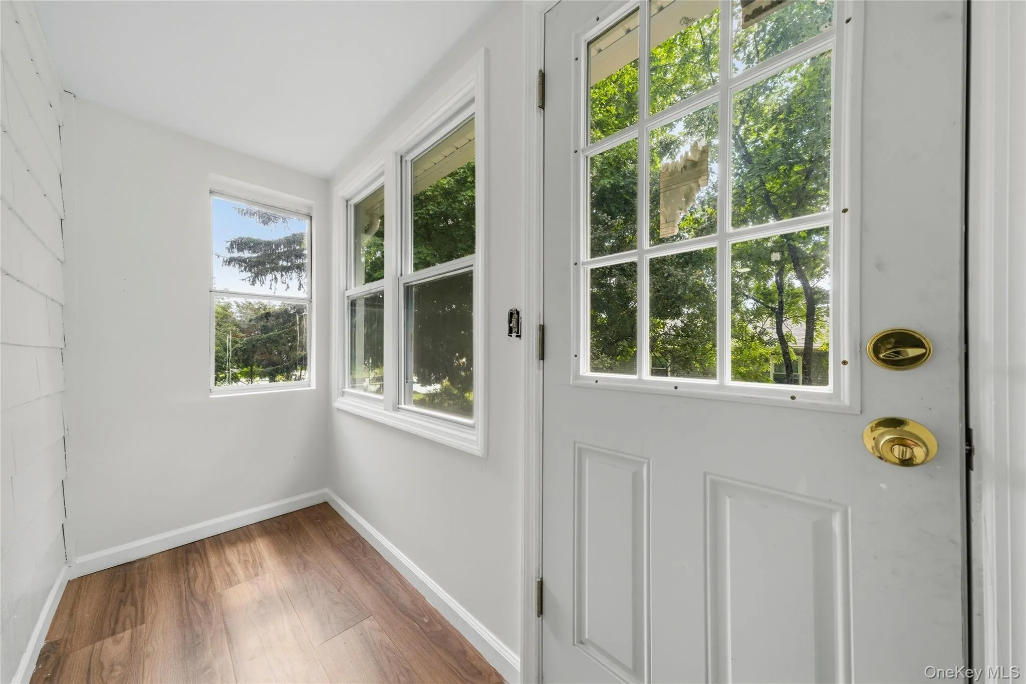 Bright mudroom entry featuring multiple windows framing lush exterior greenery, warm wood-finish flooring, white shiplap walls, and a white paneled door with brass hardware and multiple panes for natural light and ventilation Bright mudroom entry featuring multiple windows framing lush exterior greenery, warm wood-finish flooring, white shiplap walls, and a white paneled door with brass hardware and multiple panes for natural light and ventilation