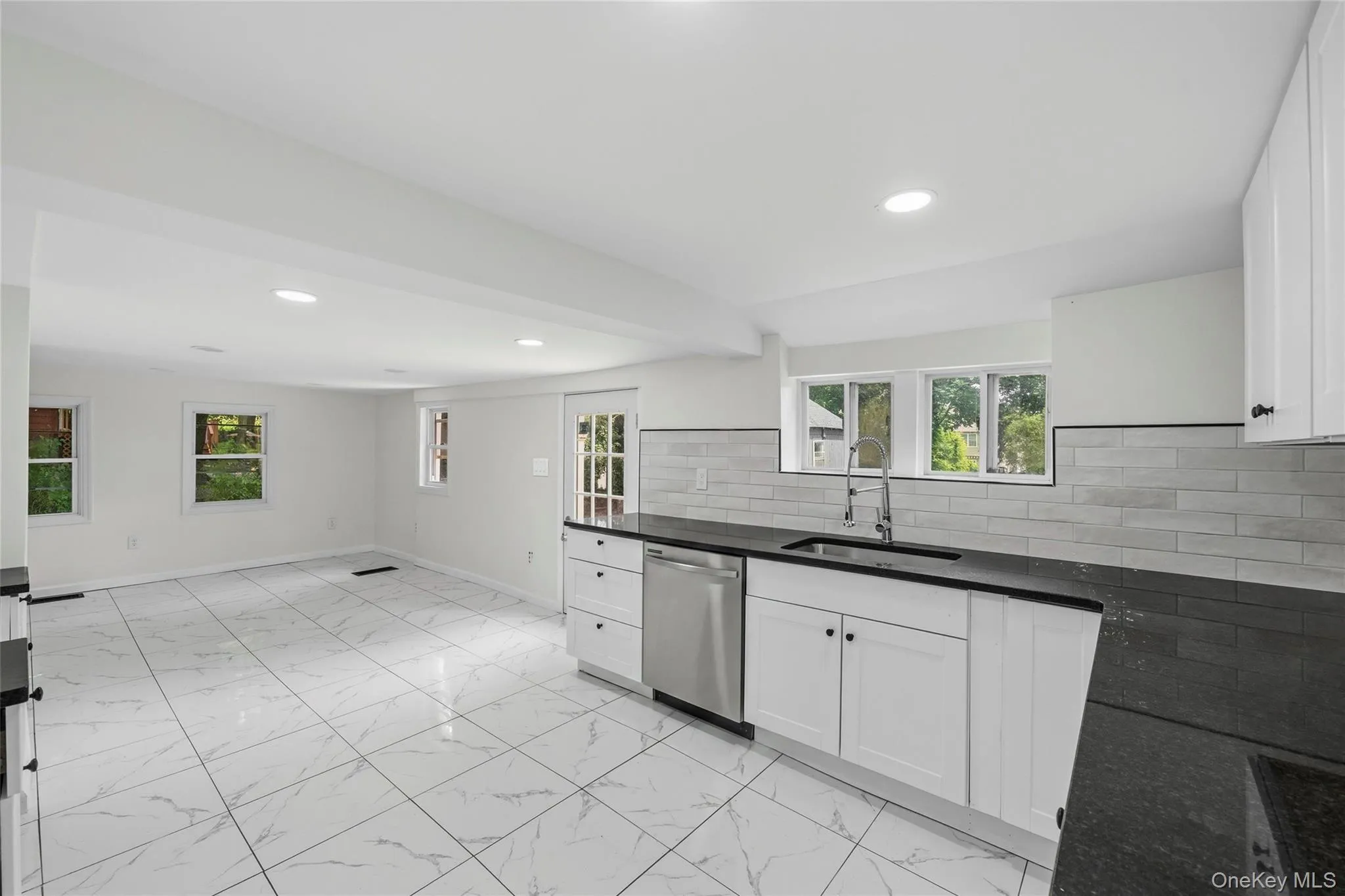Bright kitchen featuring white cabinetry, dark countertops, a stainless steel dishwasher, and a tile backsplash Bright kitchen featuring white cabinetry, dark countertops, a stainless steel dishwasher, and a tile backsplash