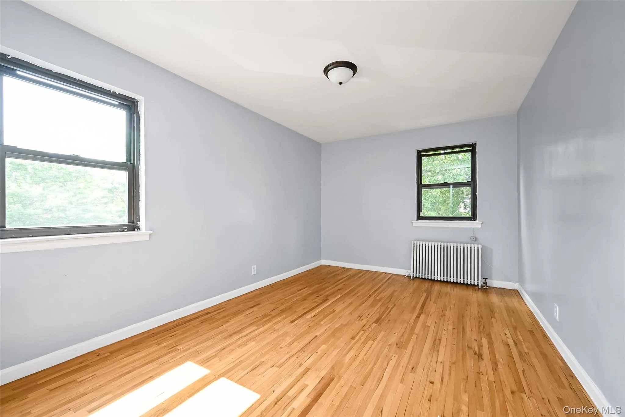 Hardwood flooring throughout, two windows with dark frames, light-colored walls, white baseboards, and a ceiling-mounted light fixture Hardwood flooring throughout, two windows with dark frames, light-colored walls, white baseboards, and a ceiling-mounted light fixture