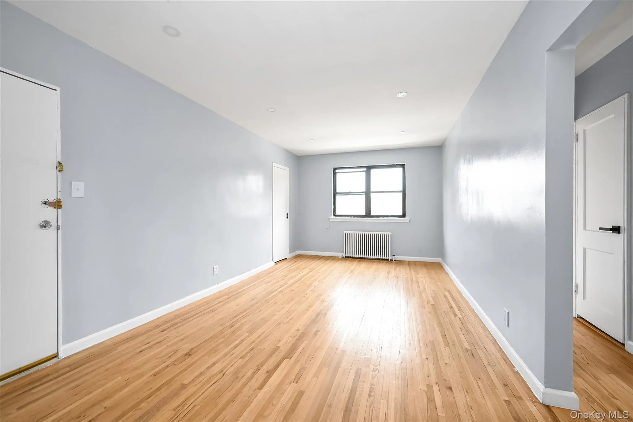 Spacious interior featuring wood-finish flooring, light blue walls, recessed ceiling lighting, a window with black trim, and a radiator Spacious interior featuring wood-finish flooring, light blue walls, recessed ceiling lighting, a window with black trim, and a radiator