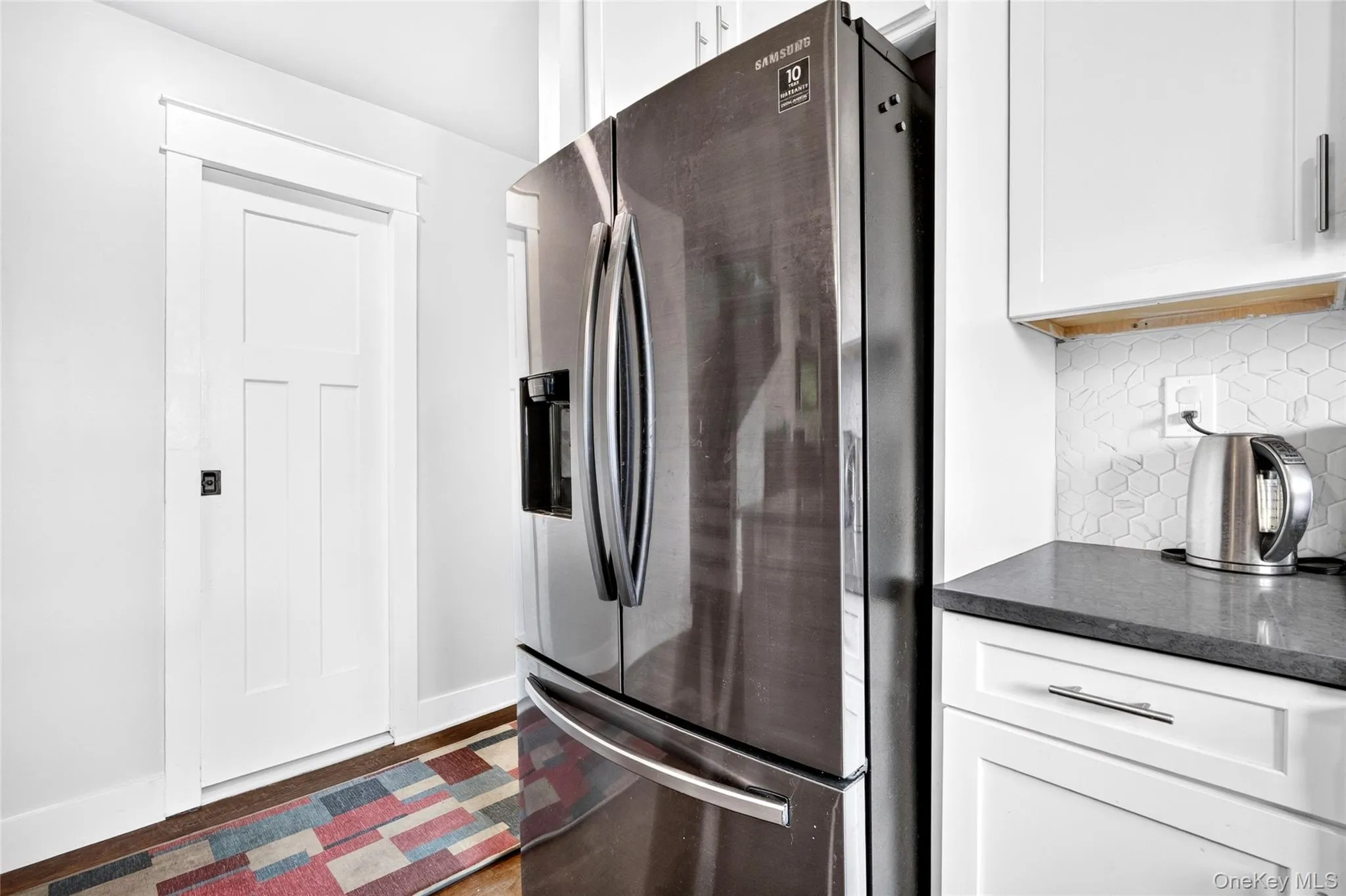 Contemporary kitchen featuring a stainless steel French door refrigerator, white shaker-style cabinetry with chrome hardware, dark countertop, and a white hexagonal tile backsplash Contemporary kitchen featuring a stainless steel French door refrigerator, white shaker-style cabinetry with chrome hardware, dark countertop, and a white hexagonal tile backsplash