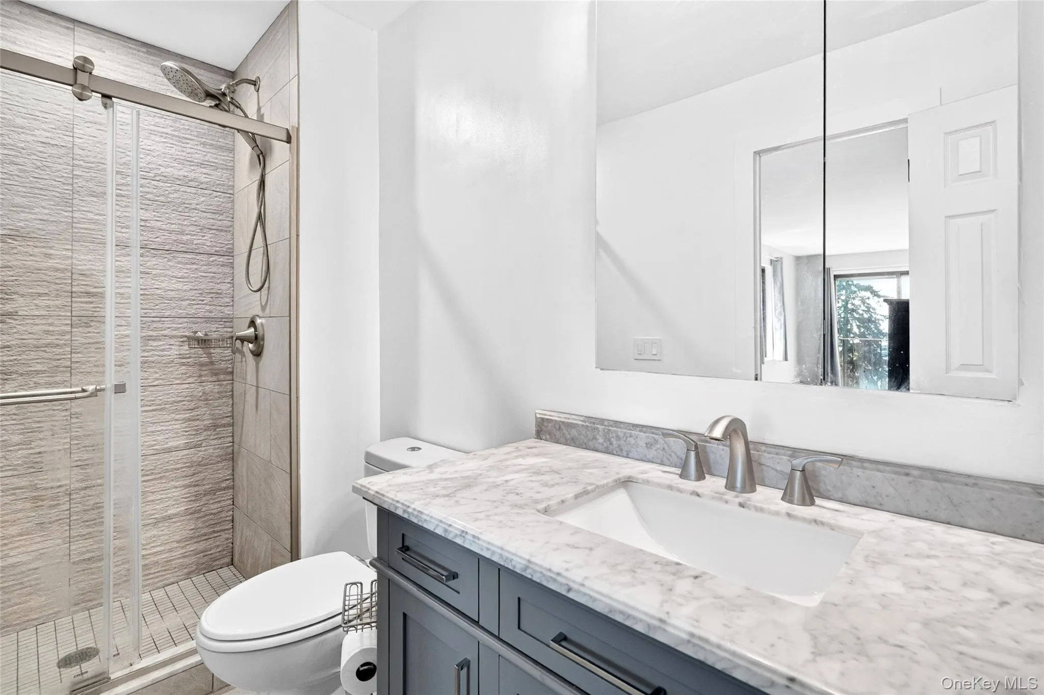 Bathroom featuring a glass-enclosed shower with textured tile, a dual-flush toilet, a vanity with a white marble-finish countertop, an undermount sink, and a mirrored medicine cabinet Bathroom featuring a glass-enclosed shower with textured tile, a dual-flush toilet, a vanity with a white marble-finish countertop, an undermount sink, and a mirrored medicine cabinet
