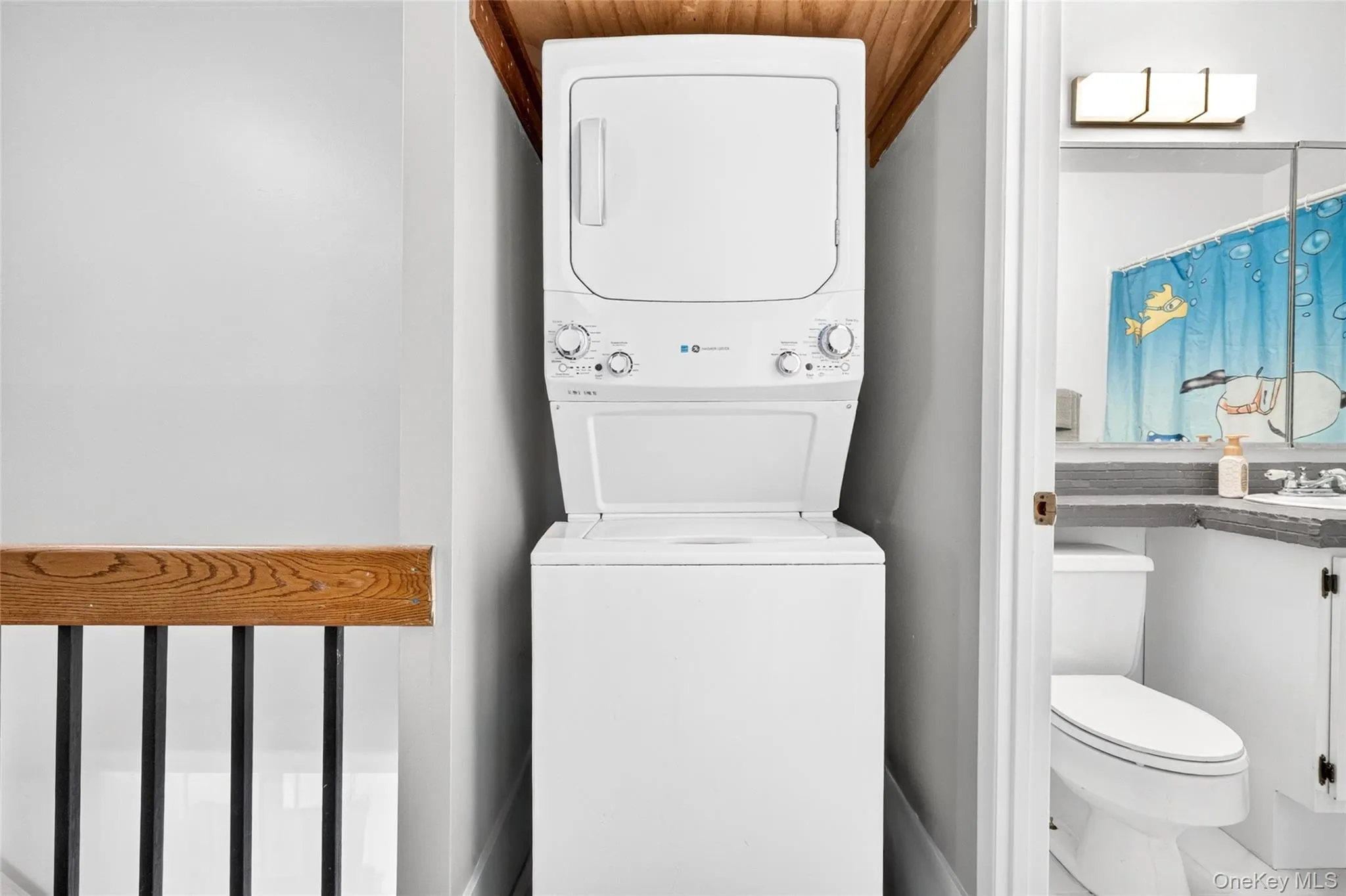 Dedicated laundry area featuring a stacked washer and dryer, complemented by a wood-finish ceiling accent and a black iron railing Dedicated laundry area featuring a stacked washer and dryer, complemented by a wood-finish ceiling accent and a black iron railing